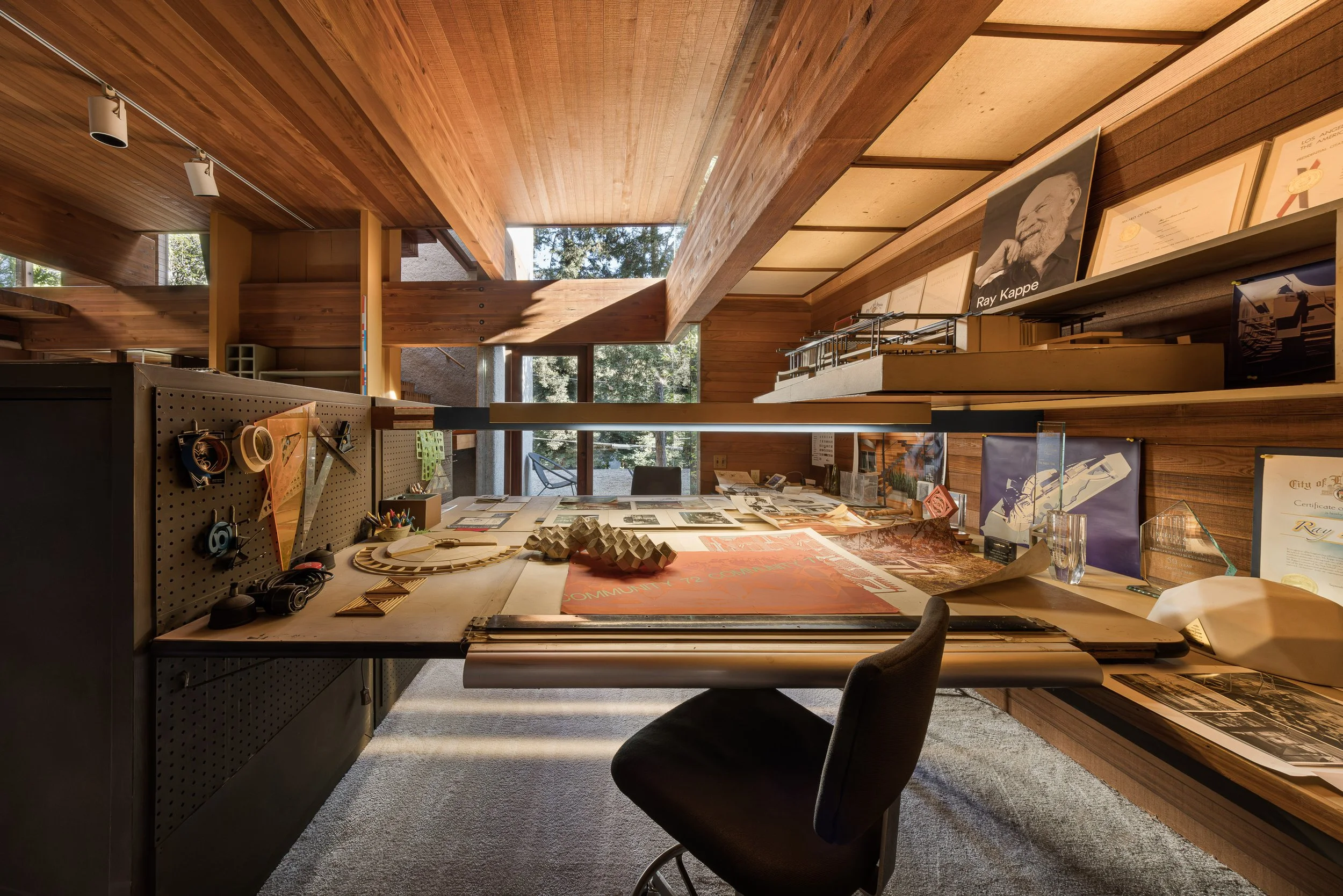 Interior of a wood-paneled office or studio space with a cluttered desk, shelves with framed photos and certificates, and large windows letting in natural light.
