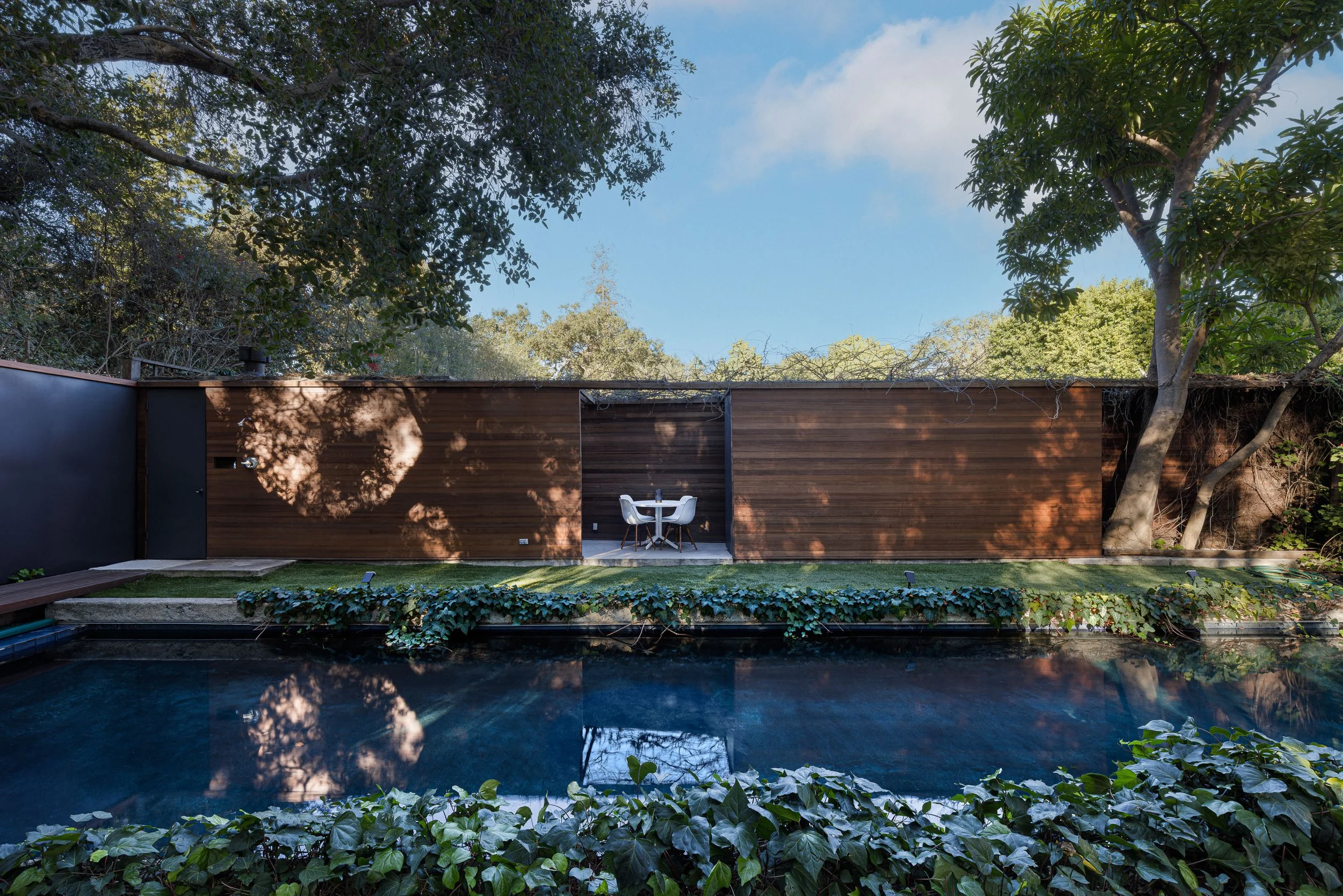 Modern backyard with a swimming pool, a wooden privacy fence, and a small patio with a table and two chairs, surrounded by green trees and landscaping.