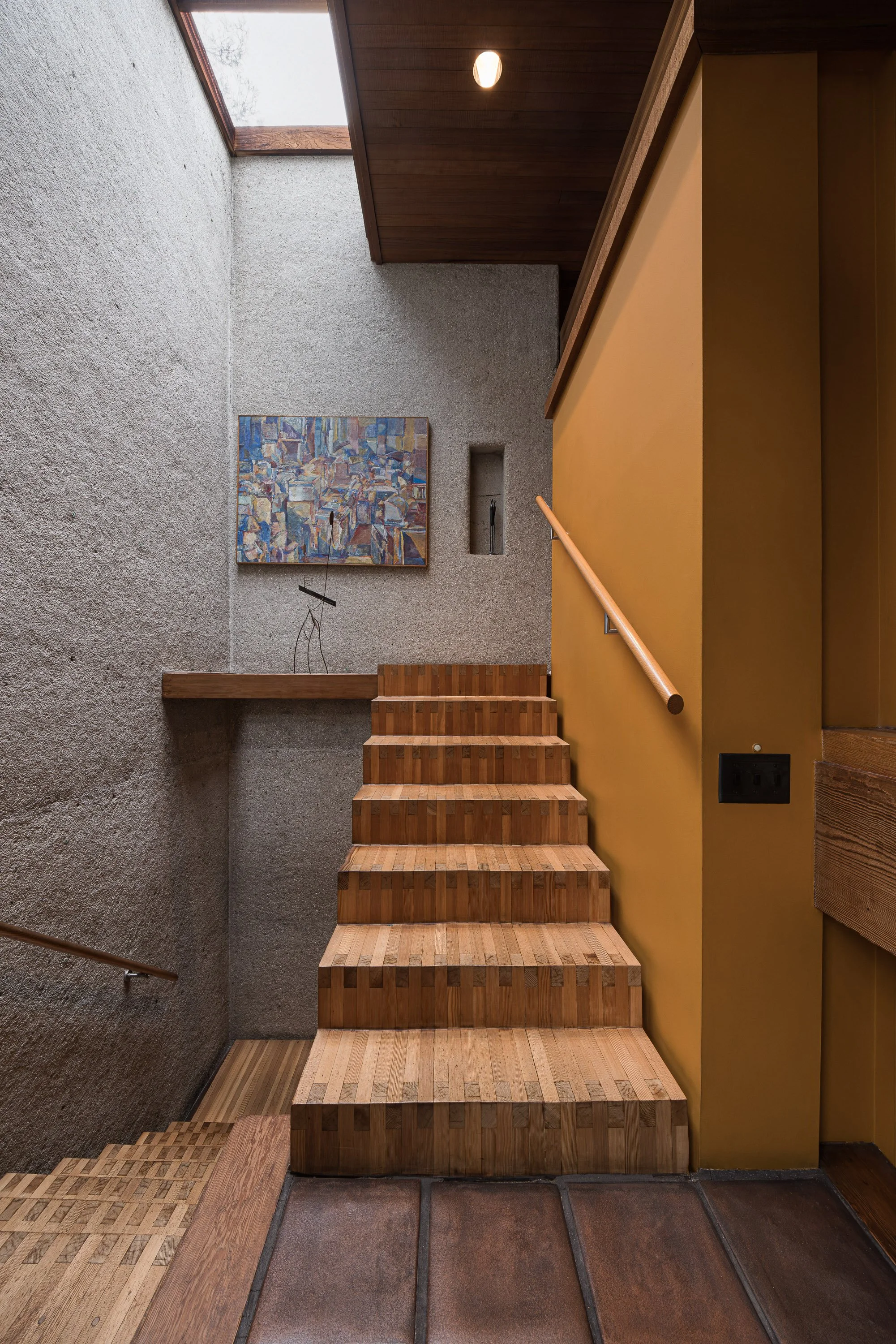 Interior staircase with wooden steps, yellow wall with power outlet, textured grey wall with a colorful abstract painting, small niche, and a skylight in the ceiling.