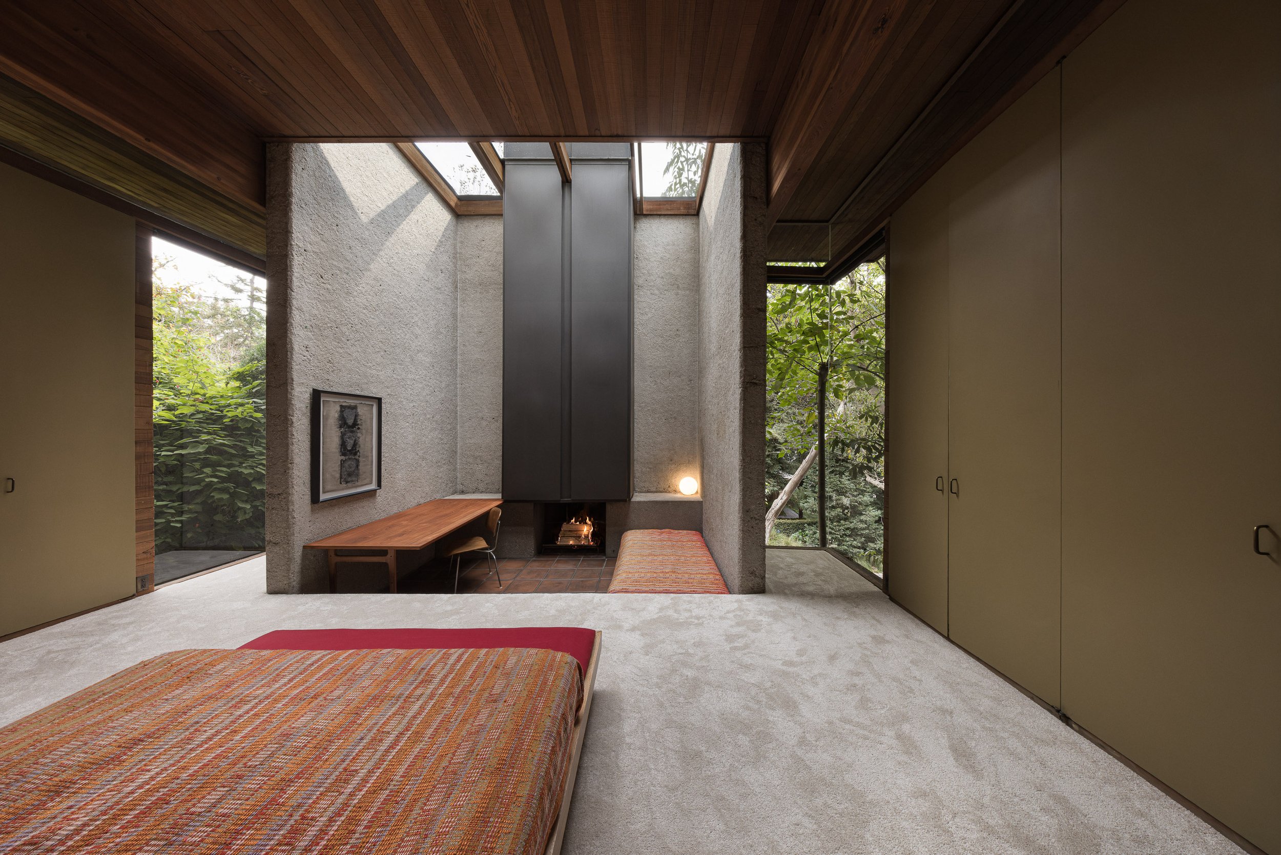 Interior of a bedroom with a low bed, wood ceiling, large windows, a fireplace, and minimalistic decor.