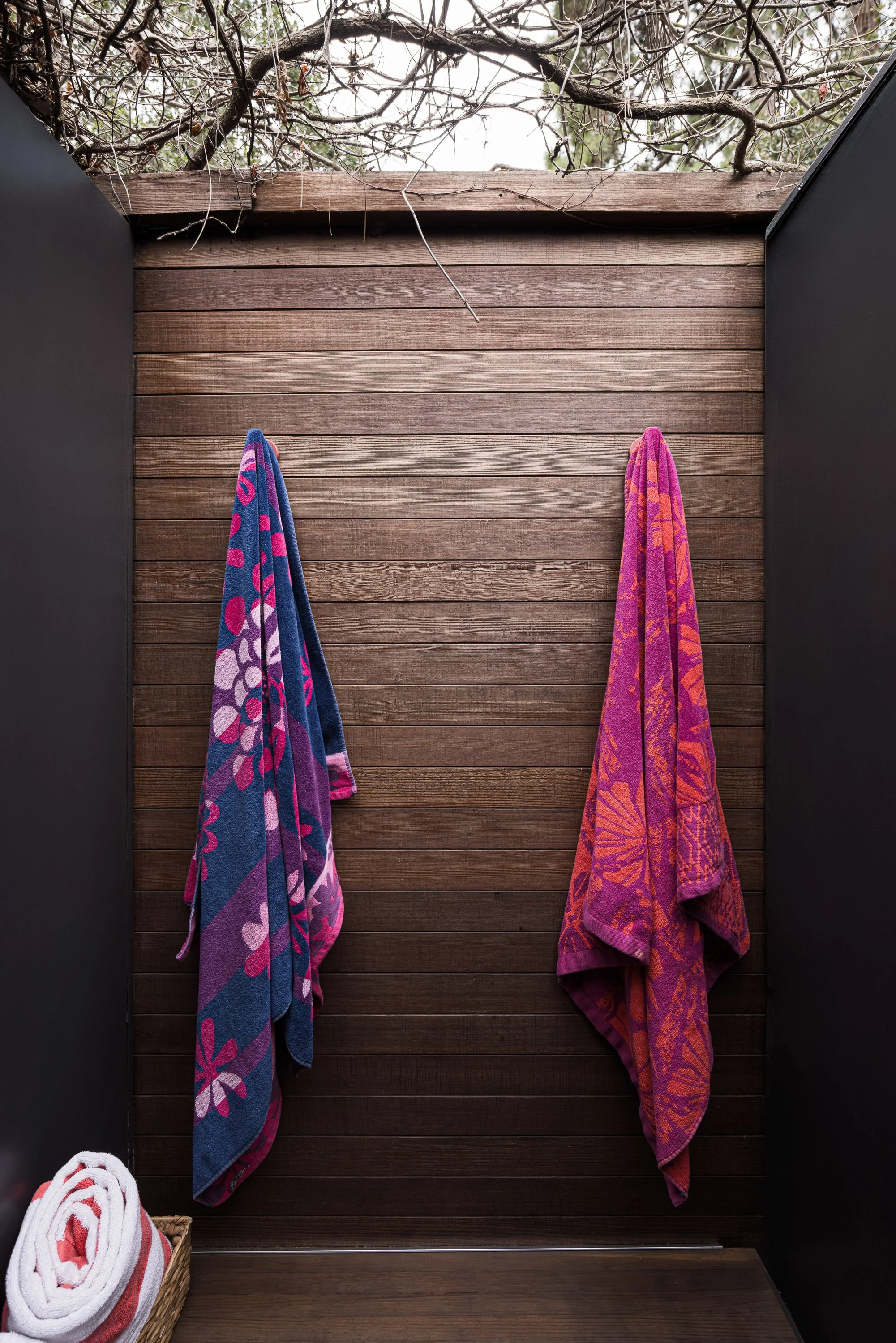 Two colorful towels hanging on hooks on a wooden wall, with a basket of rolled towels at the bottom left corner.