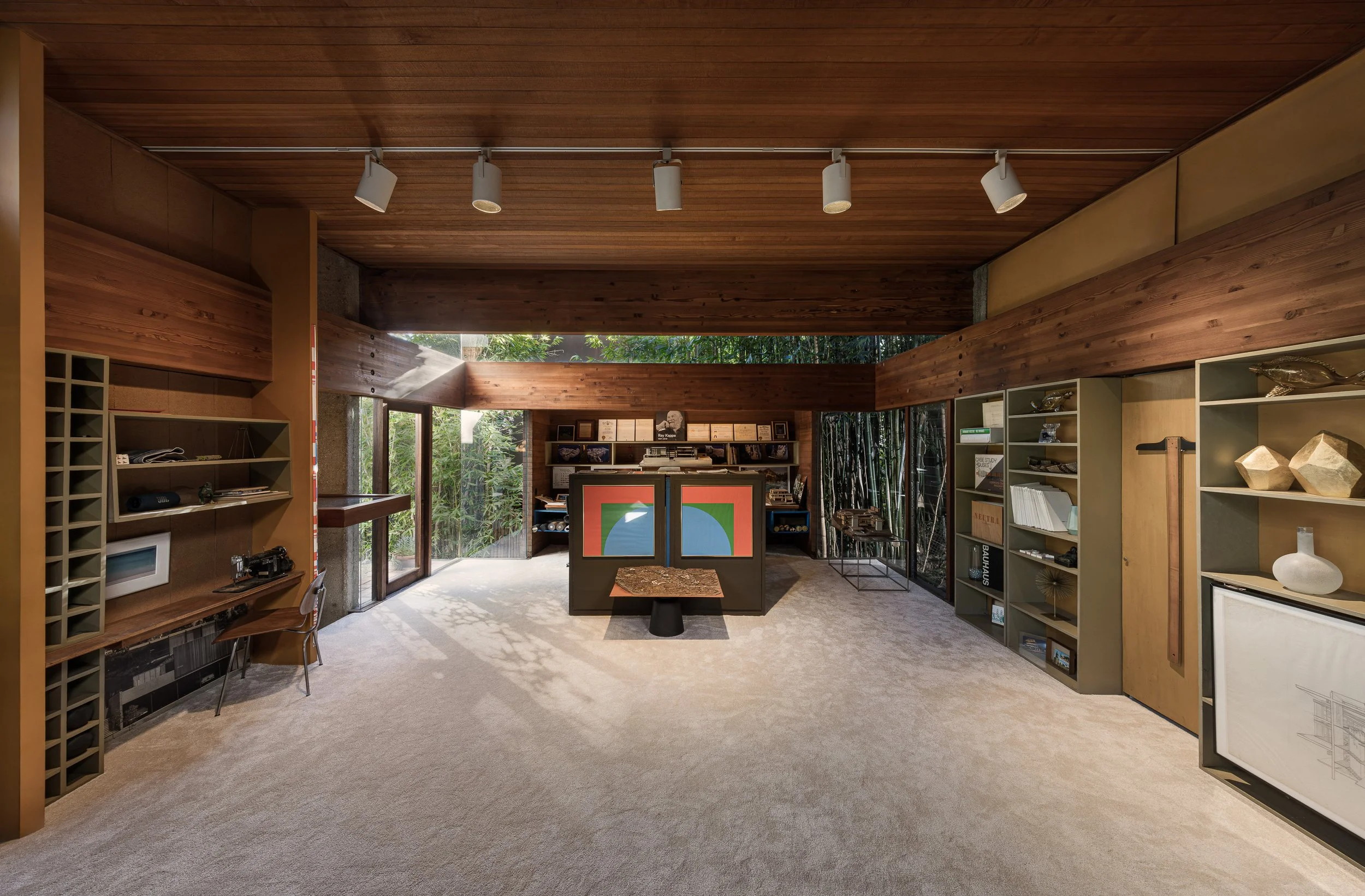 Interior of a modern room with wooden walls, ceiling, and shelves, featuring artwork, books, and decorative objects, with large windows showing greenery outside.