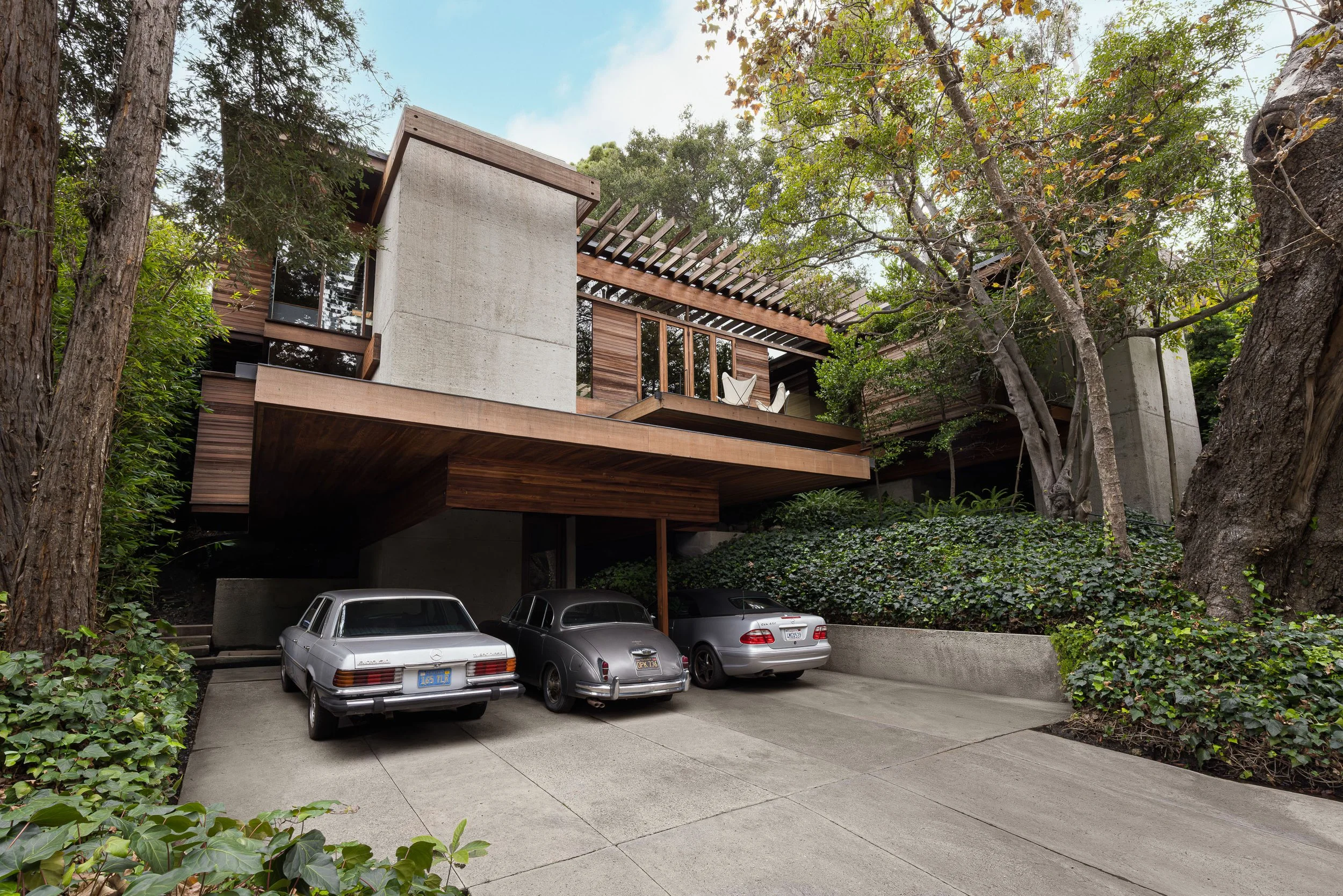 Modern multi-level house with wooden and concrete exterior, carport with three parked cars, surrounded by trees and greenery.