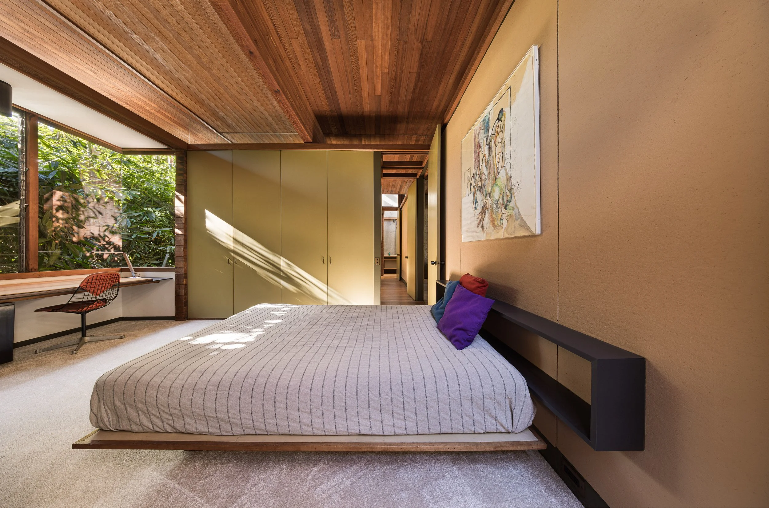Modern bedroom with large window overlooking greenery, a bed with colorful pillows, a desk with a chair, and a large closet with green doors. Wooden ceiling and beige walls.