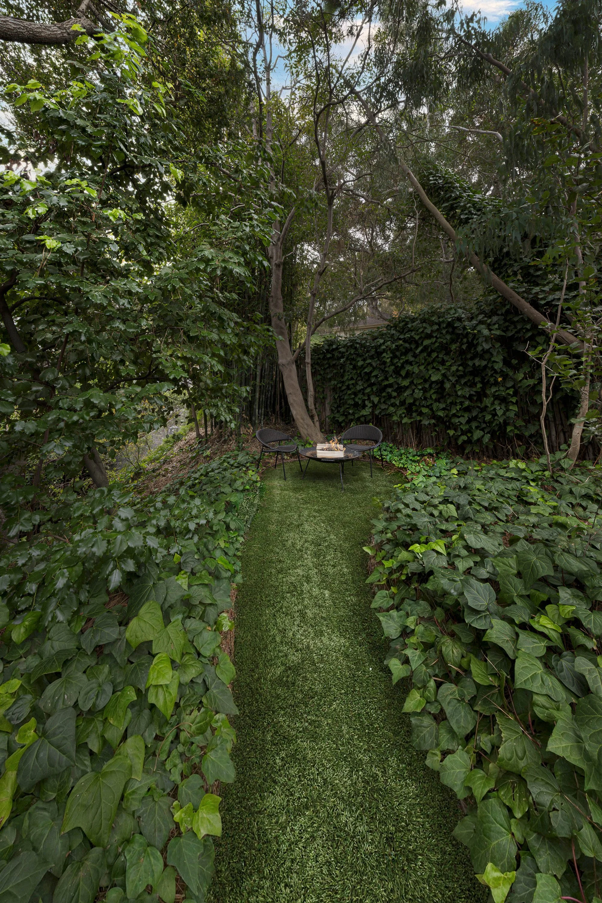 A small outdoor seating area with a round table and two black chairs on a grassy pathway surrounded by lush green ivy and trees in a private garden.