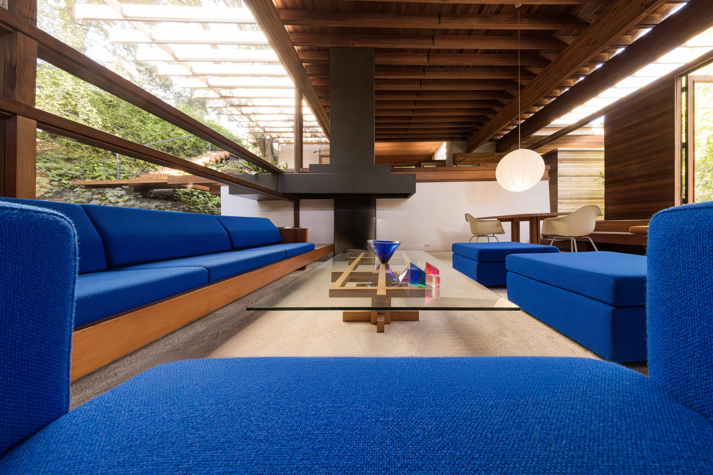 Modern living room with blue sofas, a glass coffee table, and a wooden ceiling with large windows showing greenery outside.