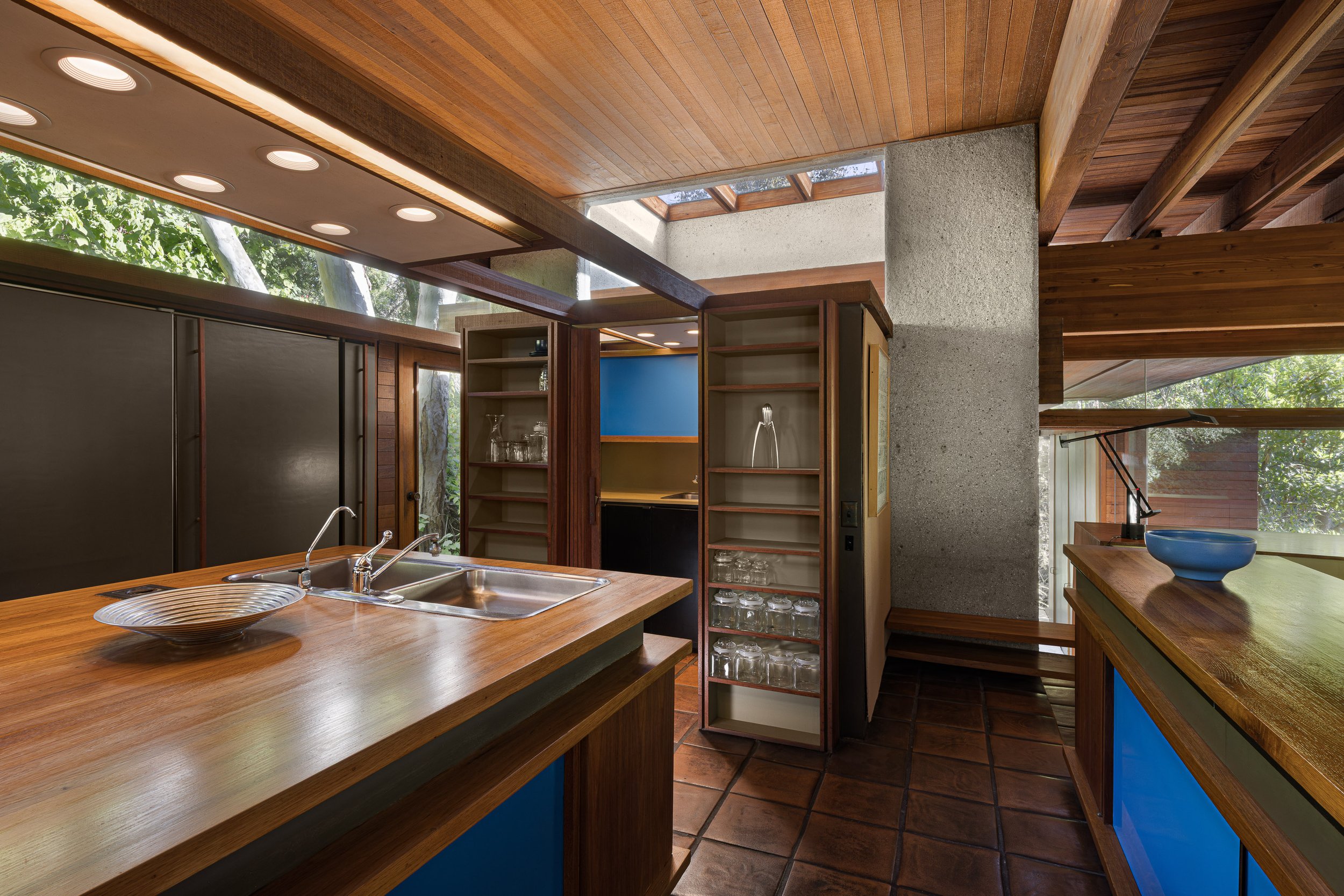 Interior view of a kitchen with wooden cabinetry, a wooden island with a sink, open shelves with glasses, and large windows letting in natural light.