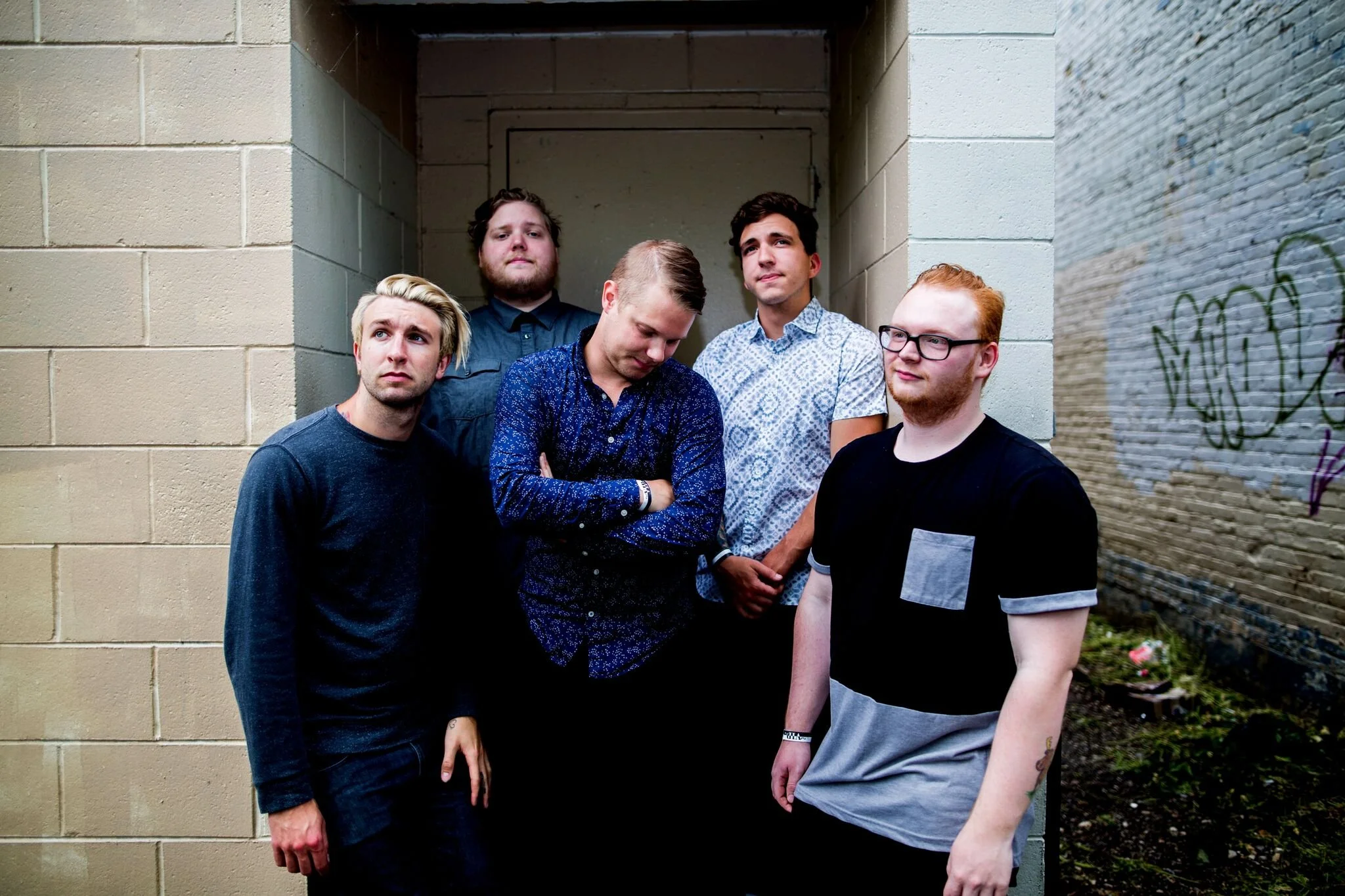Group of five men standing in a narrow alleyway with brick walls, some with graffiti, looking in different directions.