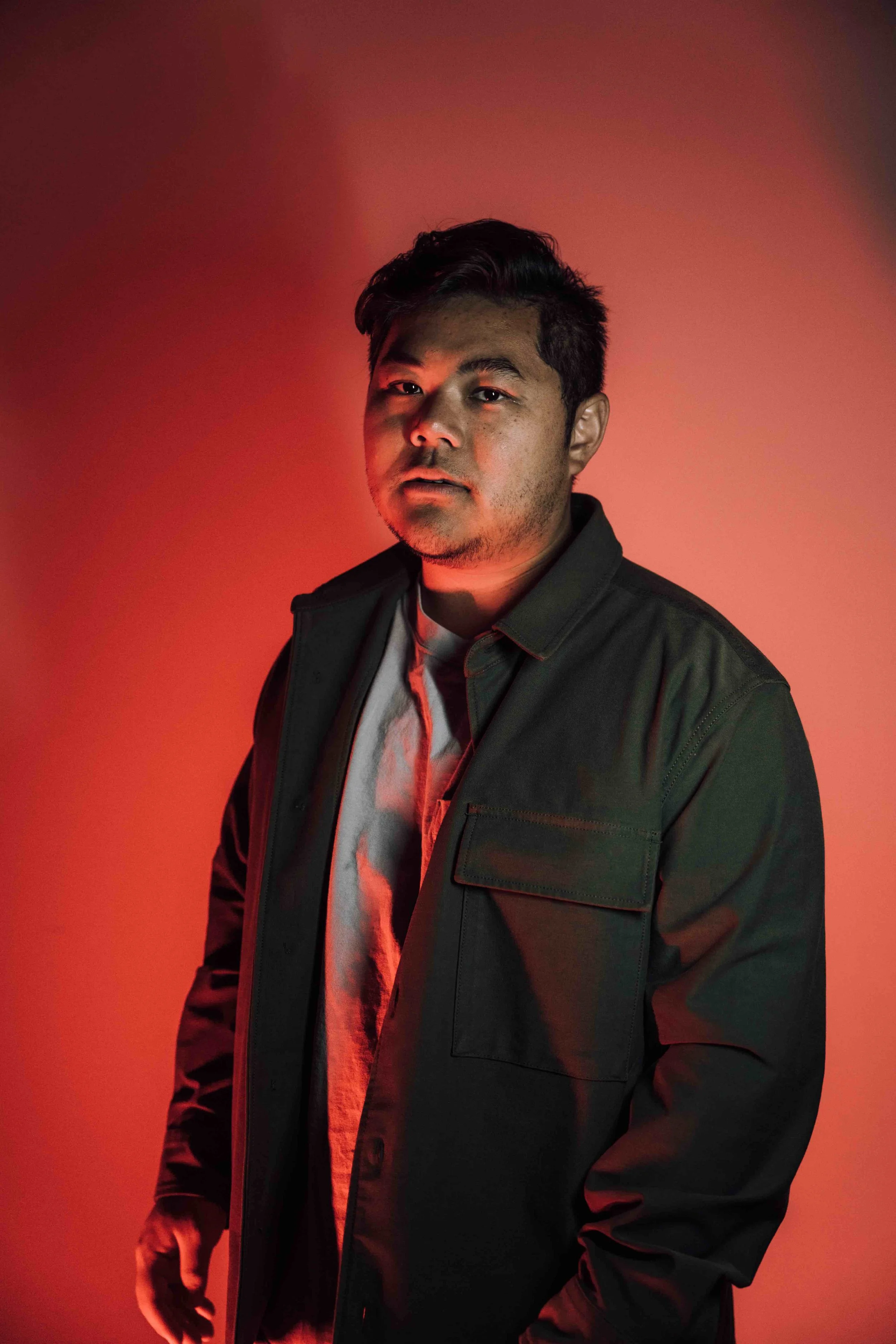 Portrait of a young man with dark hair and a slight beard, wearing a black jacket over a white shirt, standing against a red background with dramatic lighting.