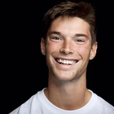 A young man with light skin, short brown hair, smiling with white teeth, wearing a white t-shirt against a black background.