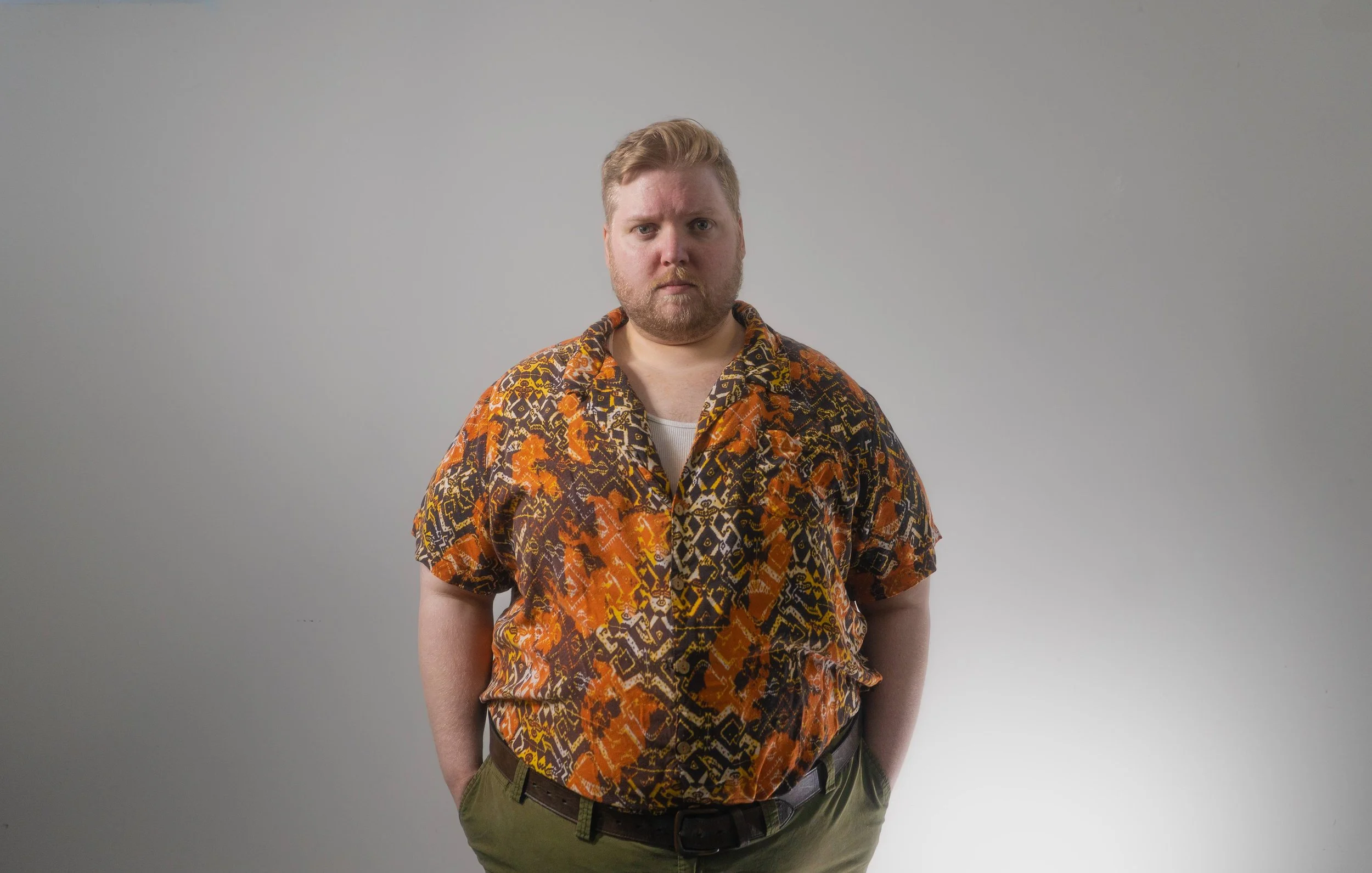 Man with a beard and short red hair wearing a colorful, patterned short-sleeve shirt and khaki pants standing against a plain light gray background.