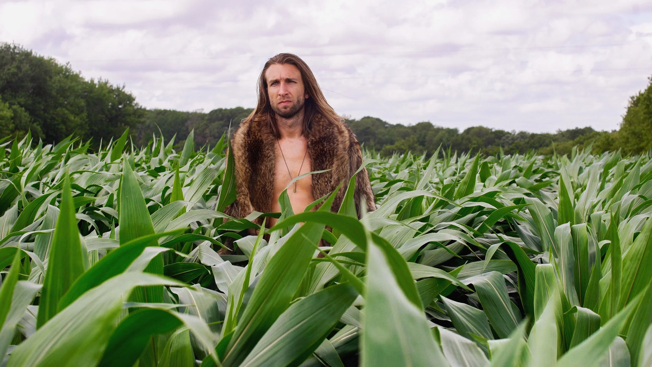 A man with long hair, wearing a fur coat open at the front, standing in a green cornfield under a cloudy sky.