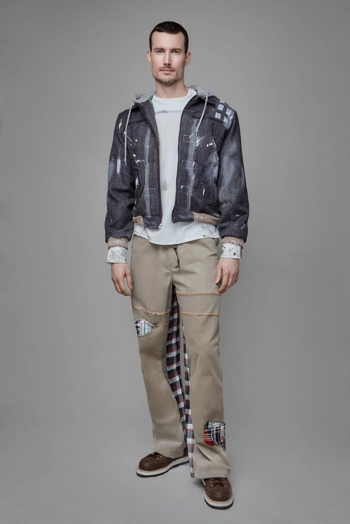 A man standing against a plain gray background, wearing a black jacket with paint splatters, a white shirt underneath, beige pants with patches and distressed details, and brown shoes.