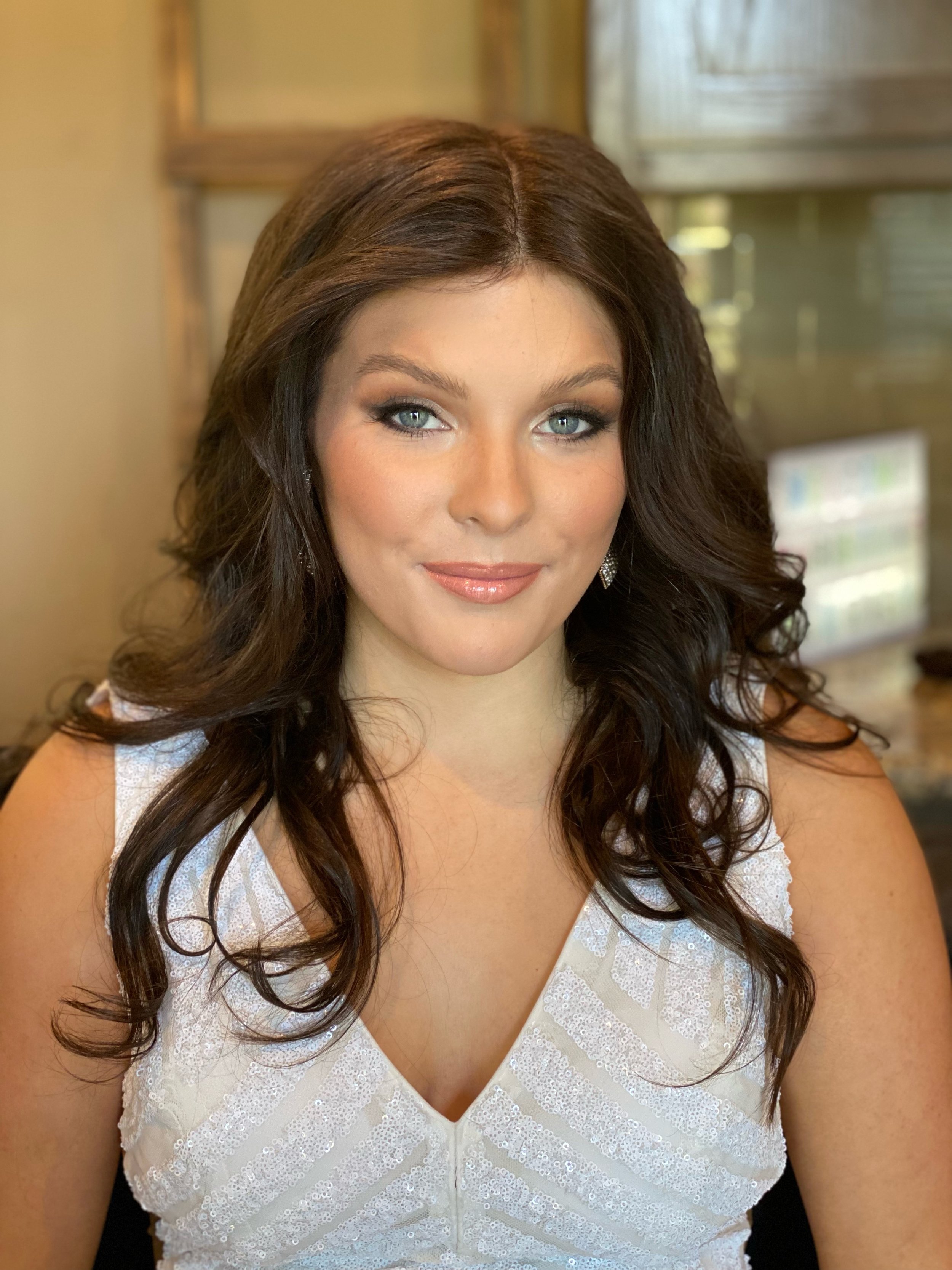 A woman with long, wavy dark brown hair, blue eyes, and makeup, wearing a white lace dress with shoulder straps and earrings, sitting indoors with a blurred background.