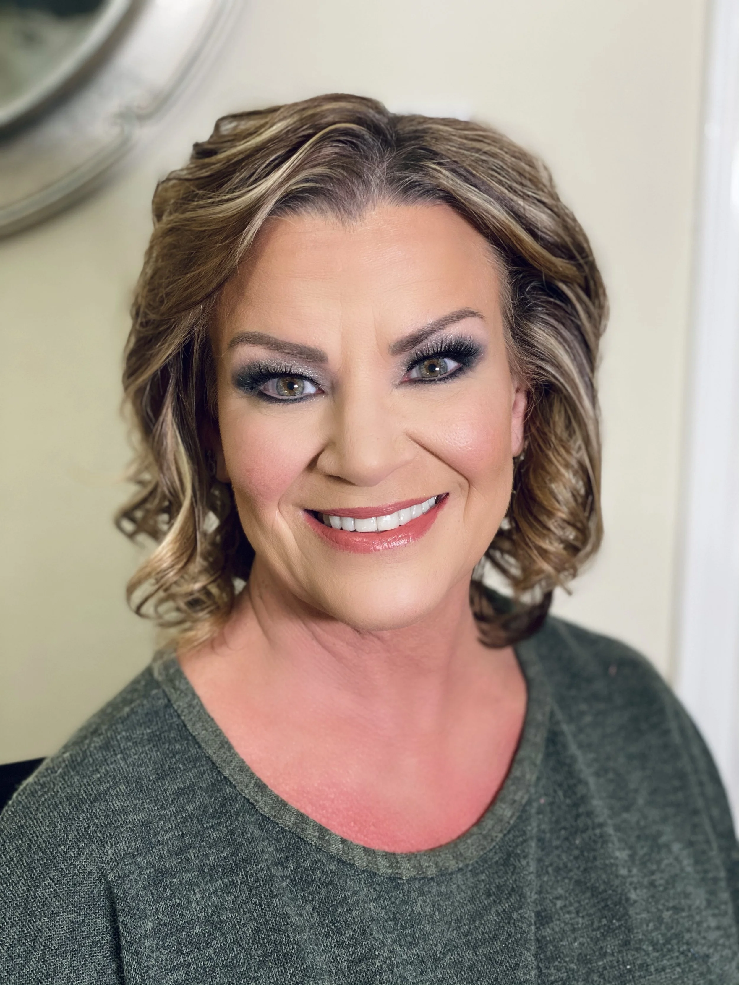 Close-up of a smiling middle-aged woman with styled brown hair, makeup, and wearing a dark green shirt, in an indoor setting.