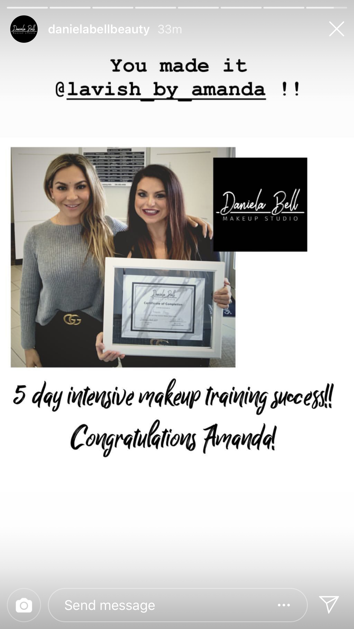 Two women smiling, one holding a framed certificate, standing in a room with a Daniels Bell Makeup Studio logo in the background. The woman on the left has blonde hair and is wearing a grey sweater and a Gucci belt, while the woman on the right has d