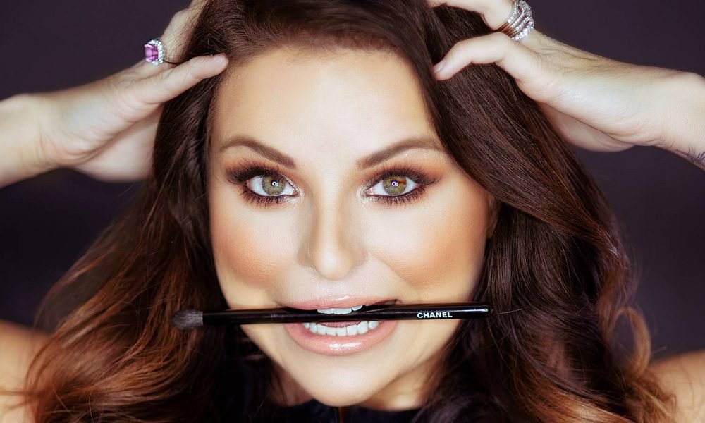 Close-up of a woman with wavy brown hair, holding her hands on her head, with a Chanel makeup brush between her teeth, against a dark background.