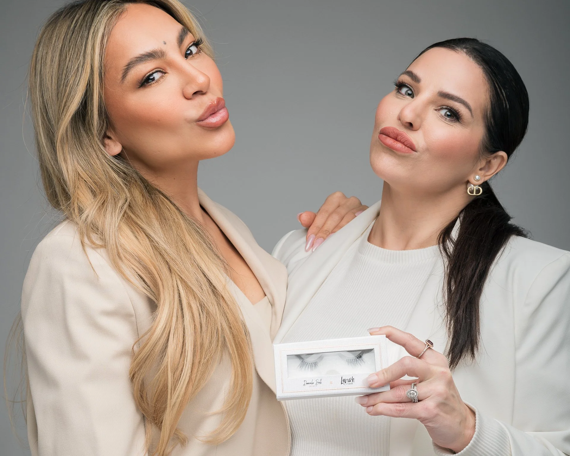 Two women dressed in beige and white outfits, with one holding a box of false eyelashes, posing together against a gray background.
