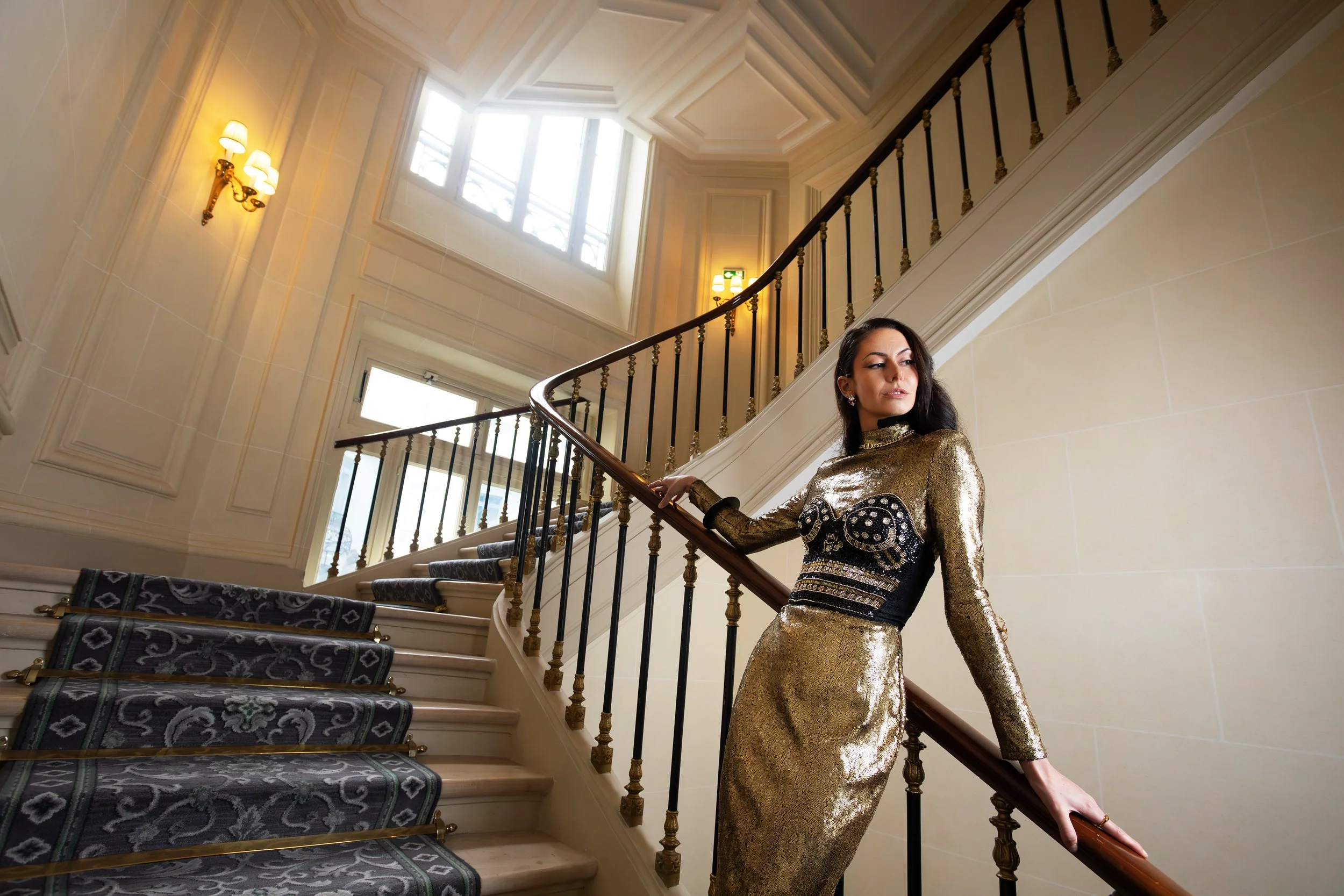 A woman in a shiny gold dress standing on a staircase inside a grand, elegant building with high ceilings and large windows.