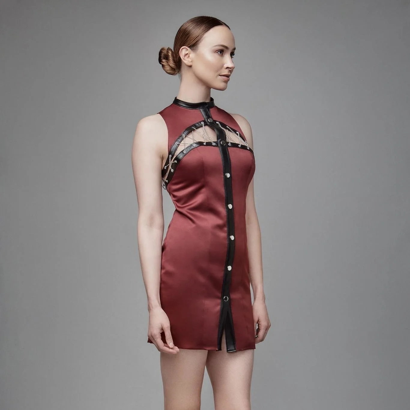 A woman wearing a red satin dress with black leather straps and silver metal details, standing against a plain gray background.