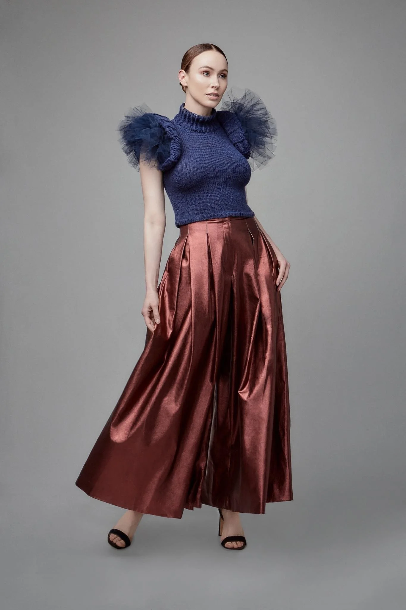 A woman wearing a navy blue sweater with tulle puffed sleeves and wide-leg, shiny bronze pants, standing against a plain gray background.