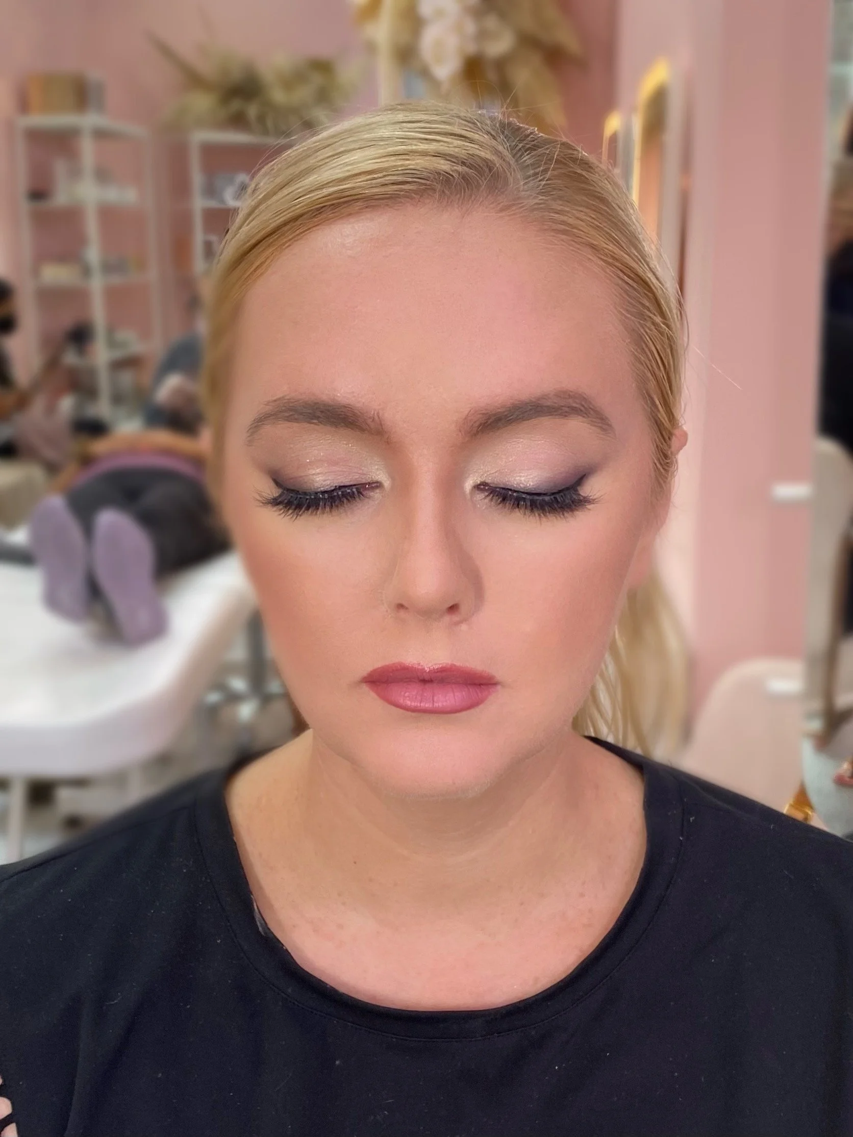 A woman with blonde hair and makeup, eyes closed, in a beauty salon with pink walls and shelves in the background.