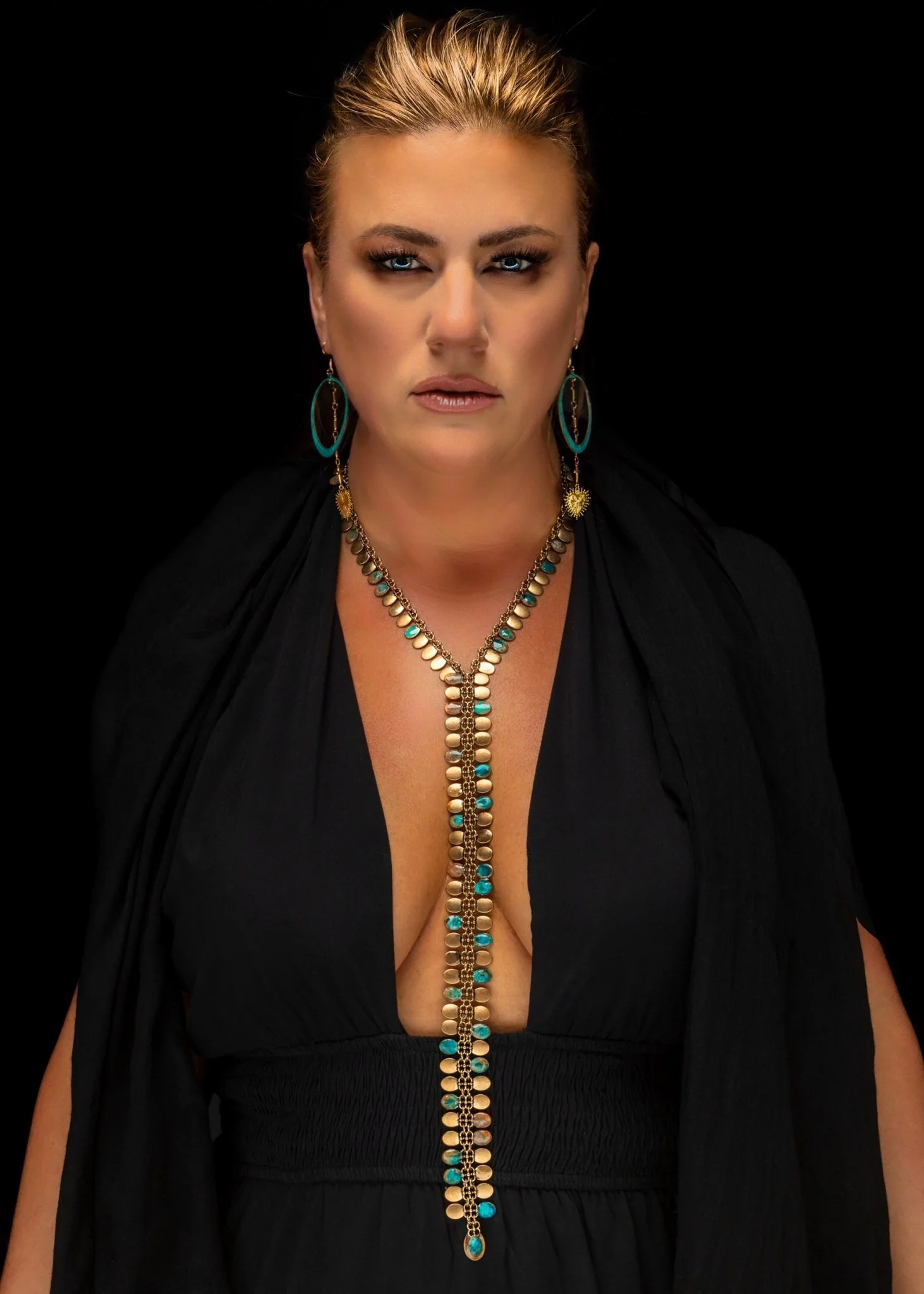 A woman with blonde hair styled back, wearing a black dress with a deep V neckline, gold and turquoise jewelry including earrings, a necklace, and a bracelet, against a black background.