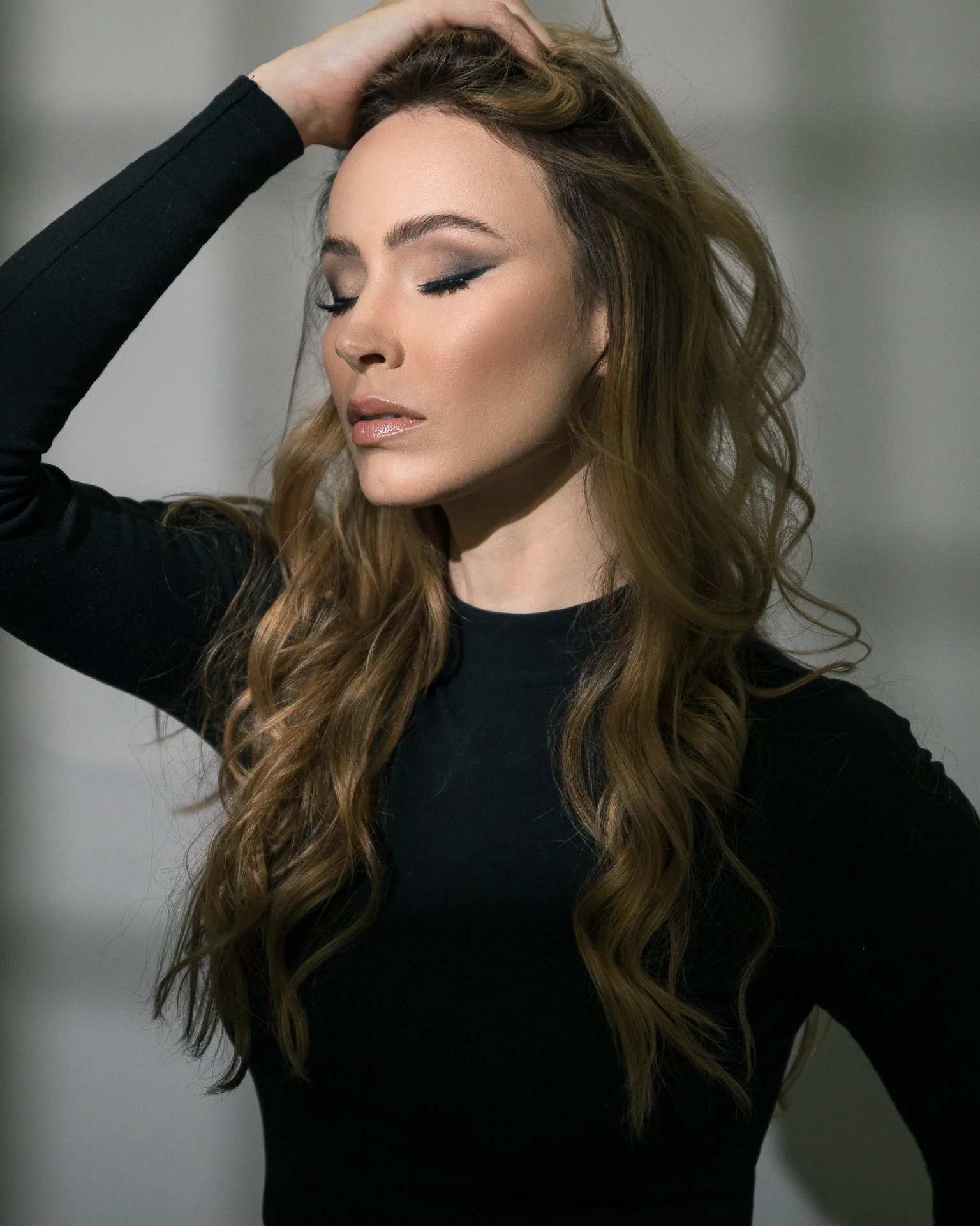 A woman with long wavy hair, wearing a black top, with her eyes closed and one hand resting on her head.