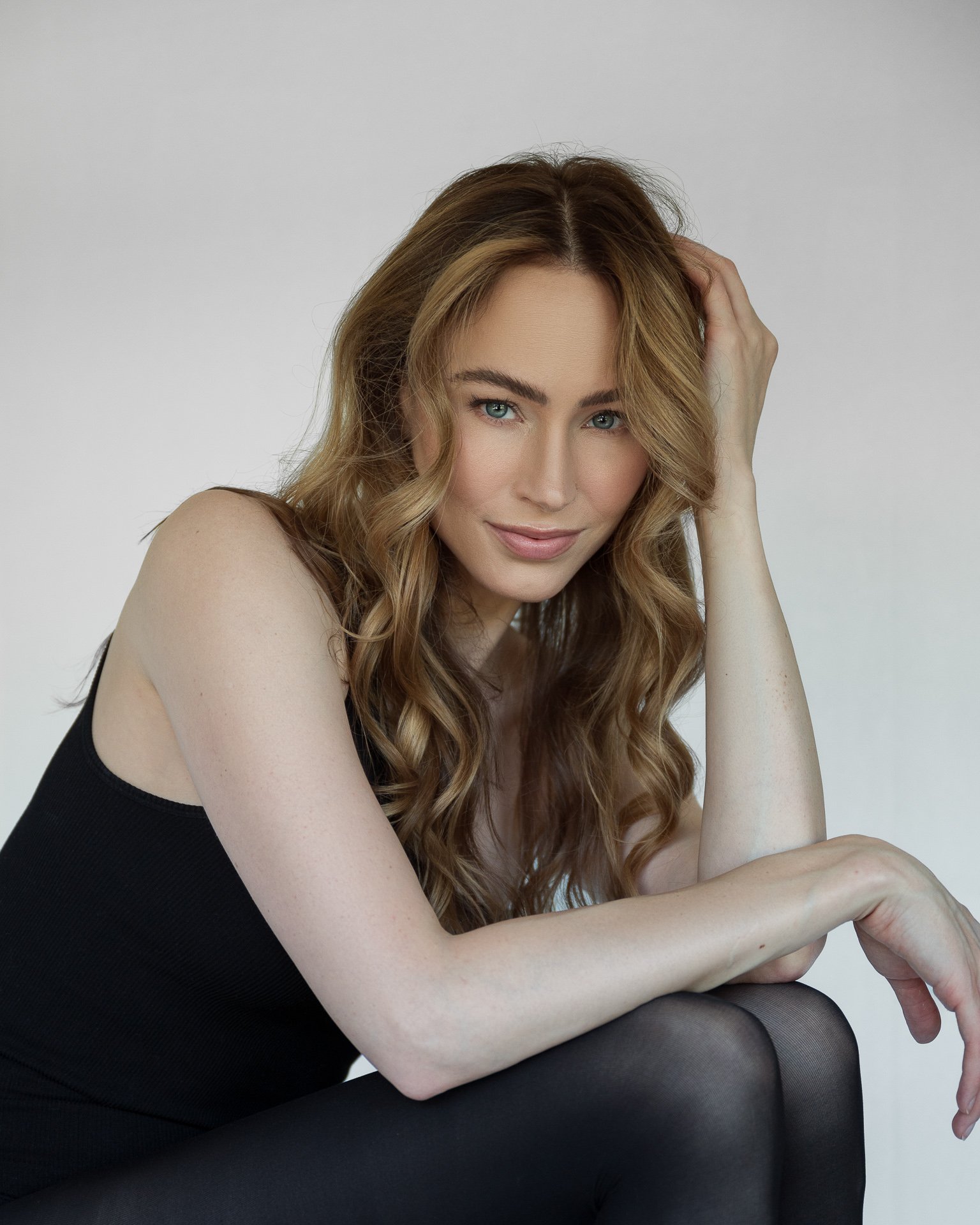 Close-up of a woman with long wavy blonde hair, blue eyes, lightly smiling, wearing a black sleeveless top, sitting with her arm resting on her knee, against a plain light background.