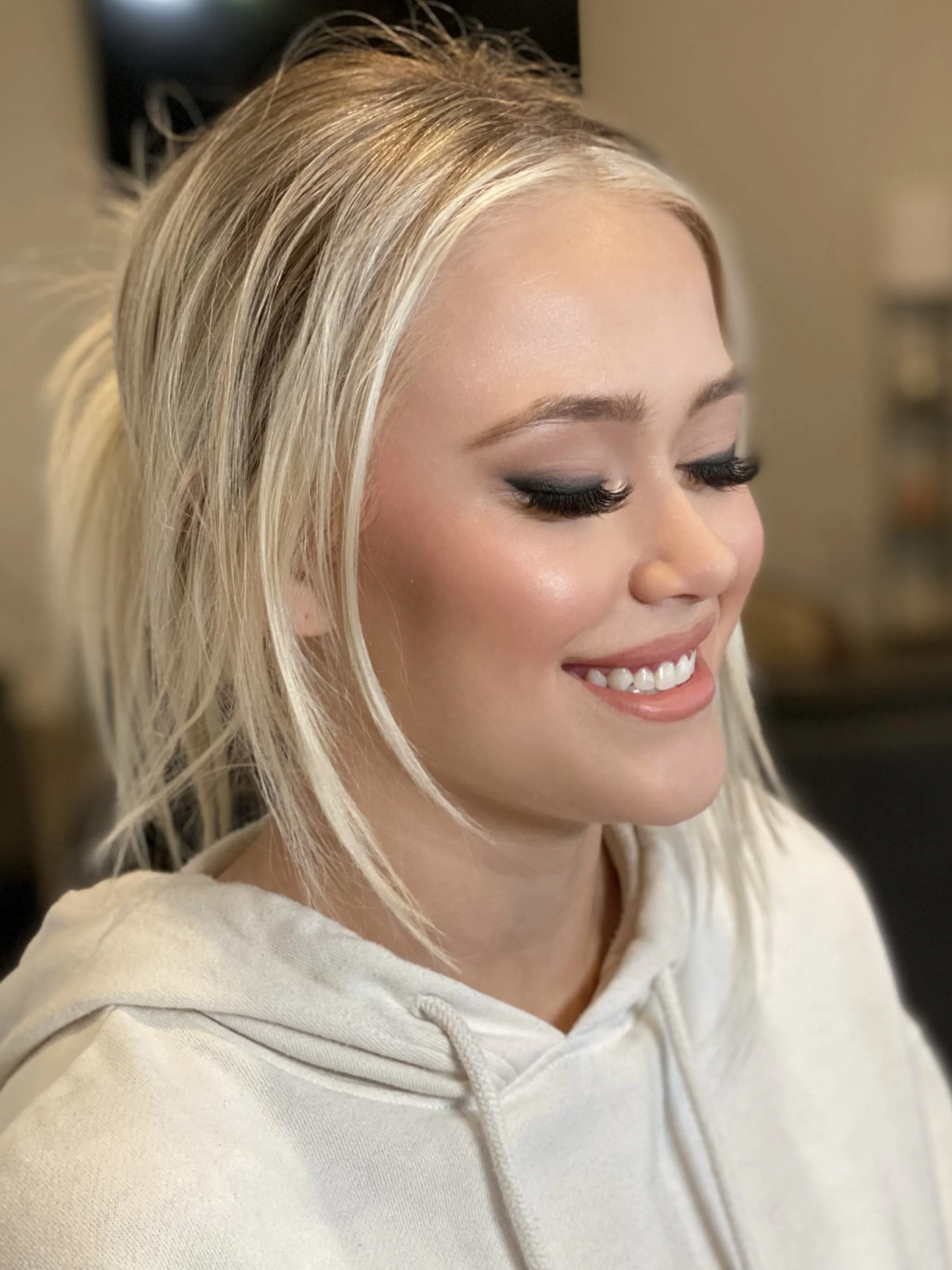 A young woman with blonde hair in a tousled style, wearing makeup with dark eyeshadow, long eyelashes, and a natural pink lip, smiling with her eyes closed, wearing a light-colored hoodie.