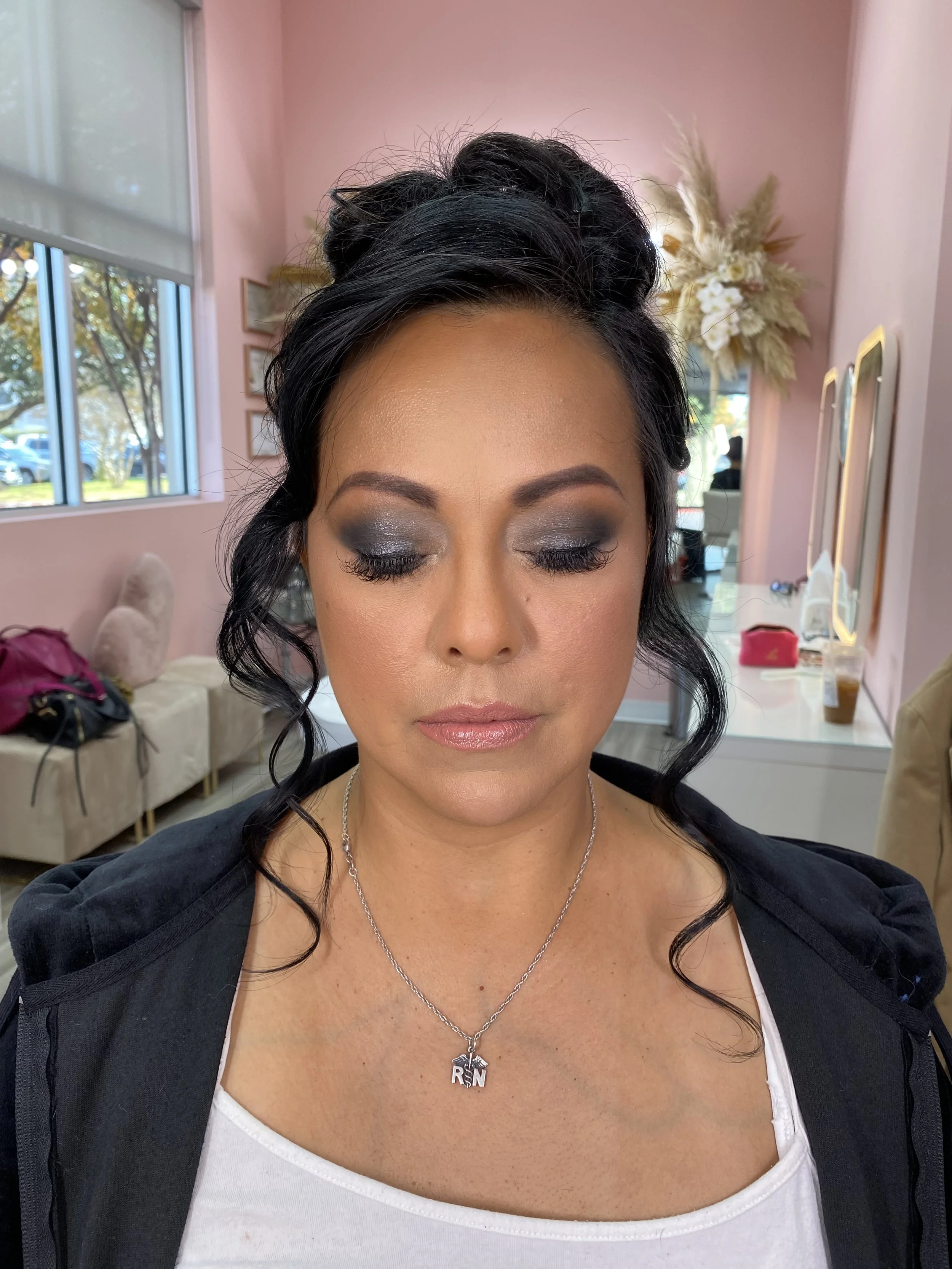 A woman with dark, curly hair and makeup showing silver eyeshadow and pink lipstick, wearing a white top and a necklace with a pendant that reads 'RN', sitting in a pink room with a vanity mirror, flowers, and various items on the dresser.