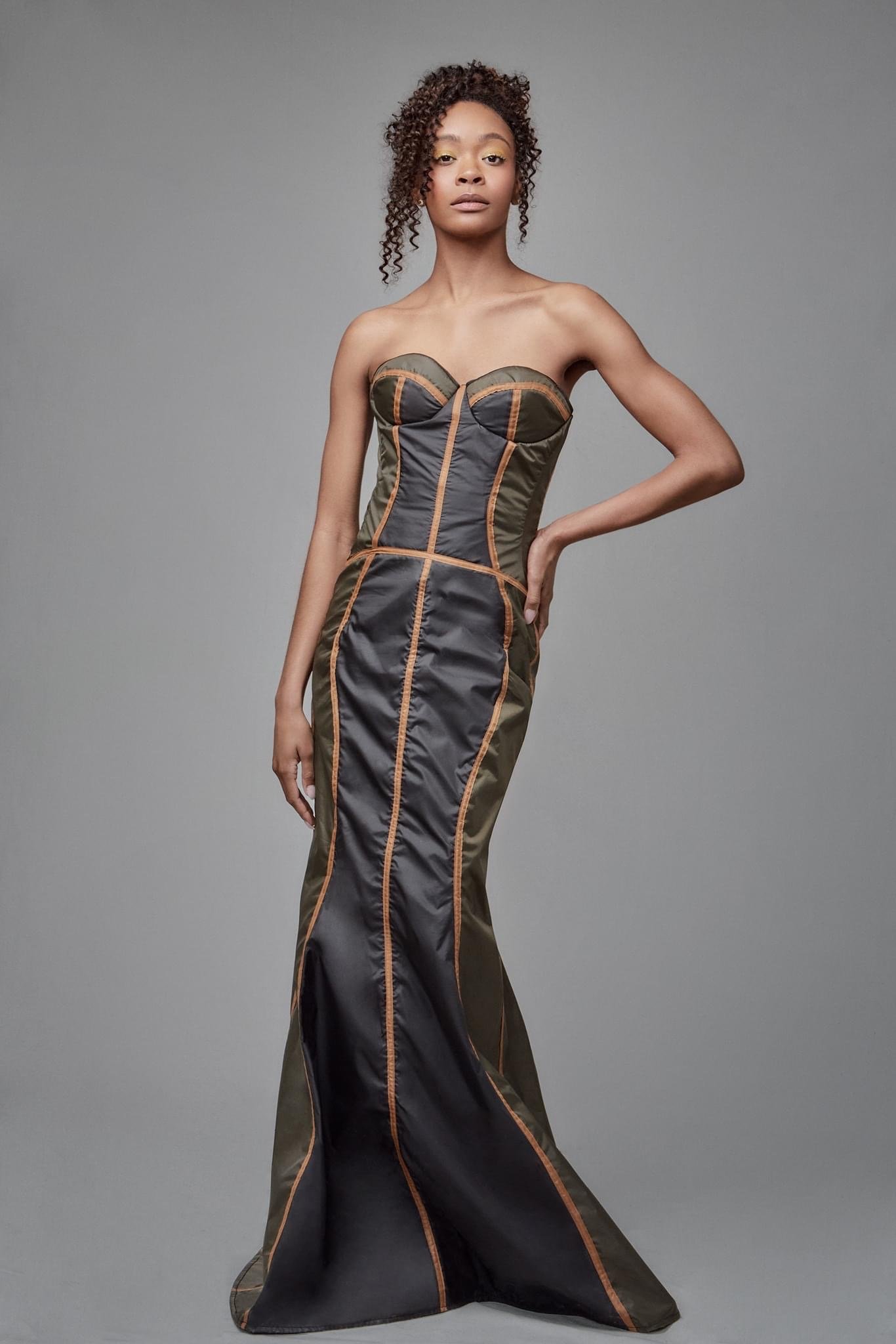 A woman wearing a strapless, black and green gown with orange piping, standing against a neutral gray background.