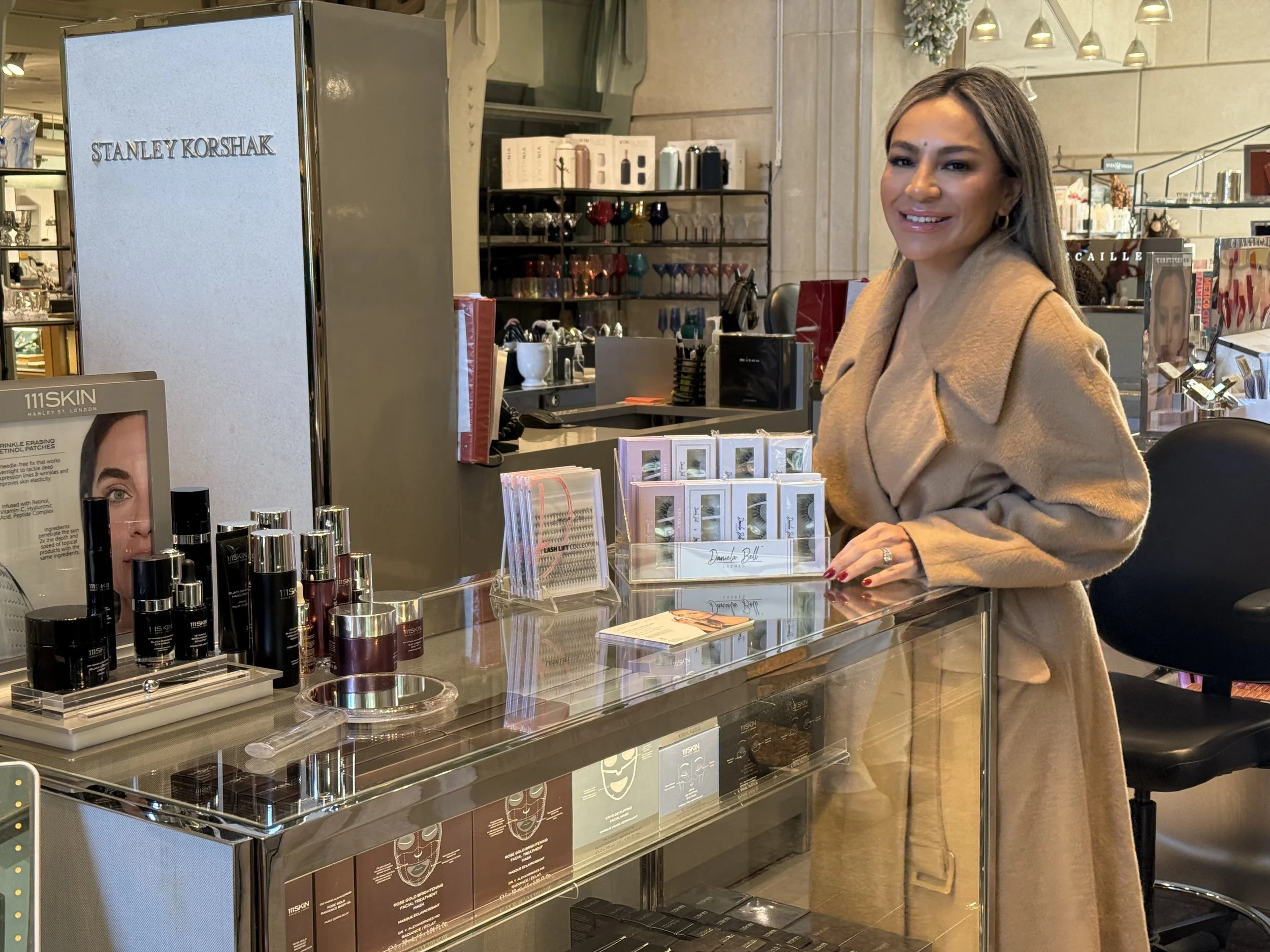 A woman with long hair and a beige coat smiling at the camera while standing at a cosmetics counter in a store, with skincare and makeup products displayed on the glass surface in front of her.