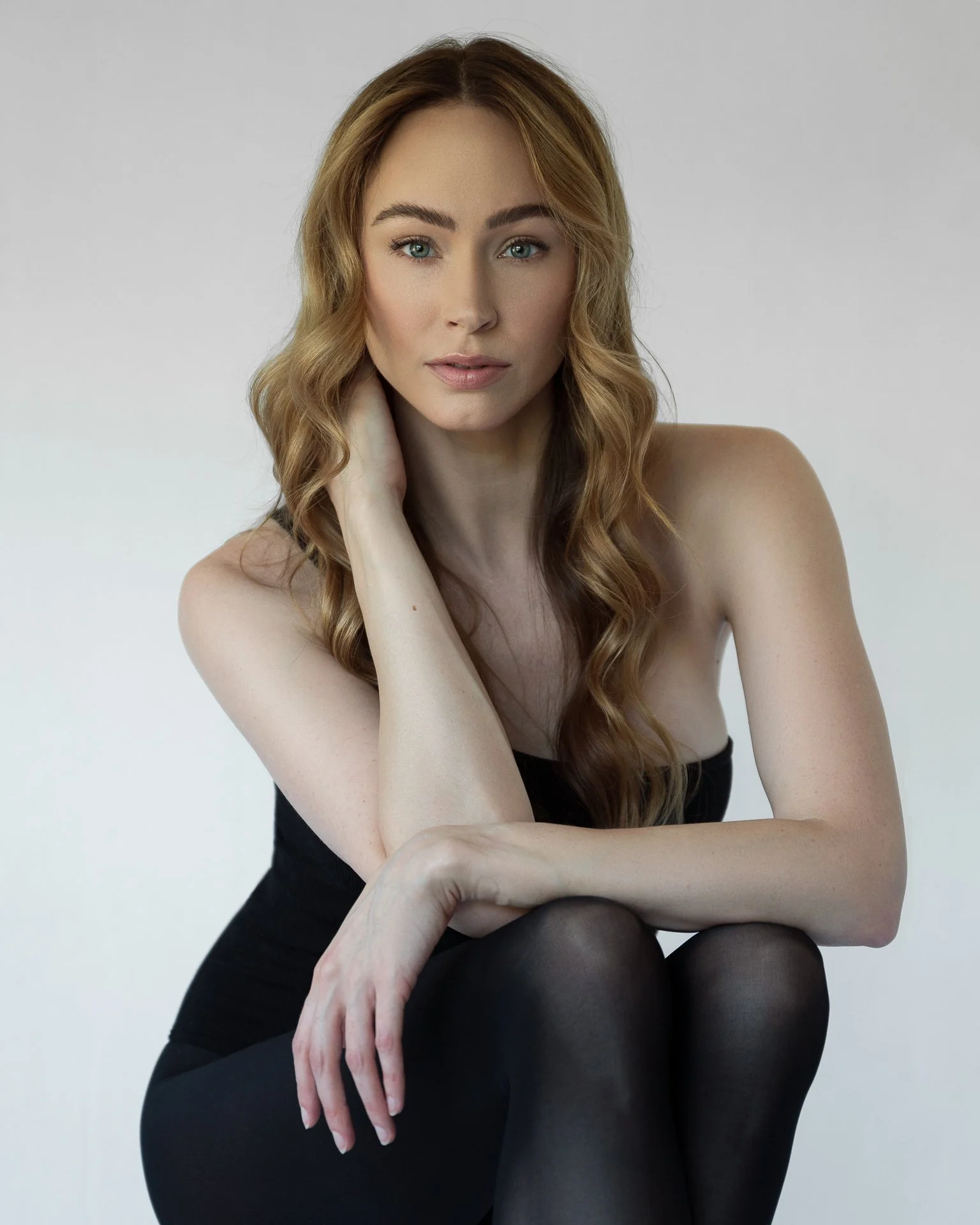 Portrait of a young woman with long wavy blond hair, blue eyes, wearing a black strapless top and black tights, sitting against a plain light gray background.