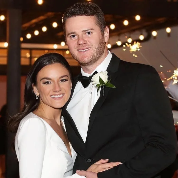A smiling couple at a formal event, the woman in a white dress and the man in a black tuxedo with a white boutonniere, standing close together.