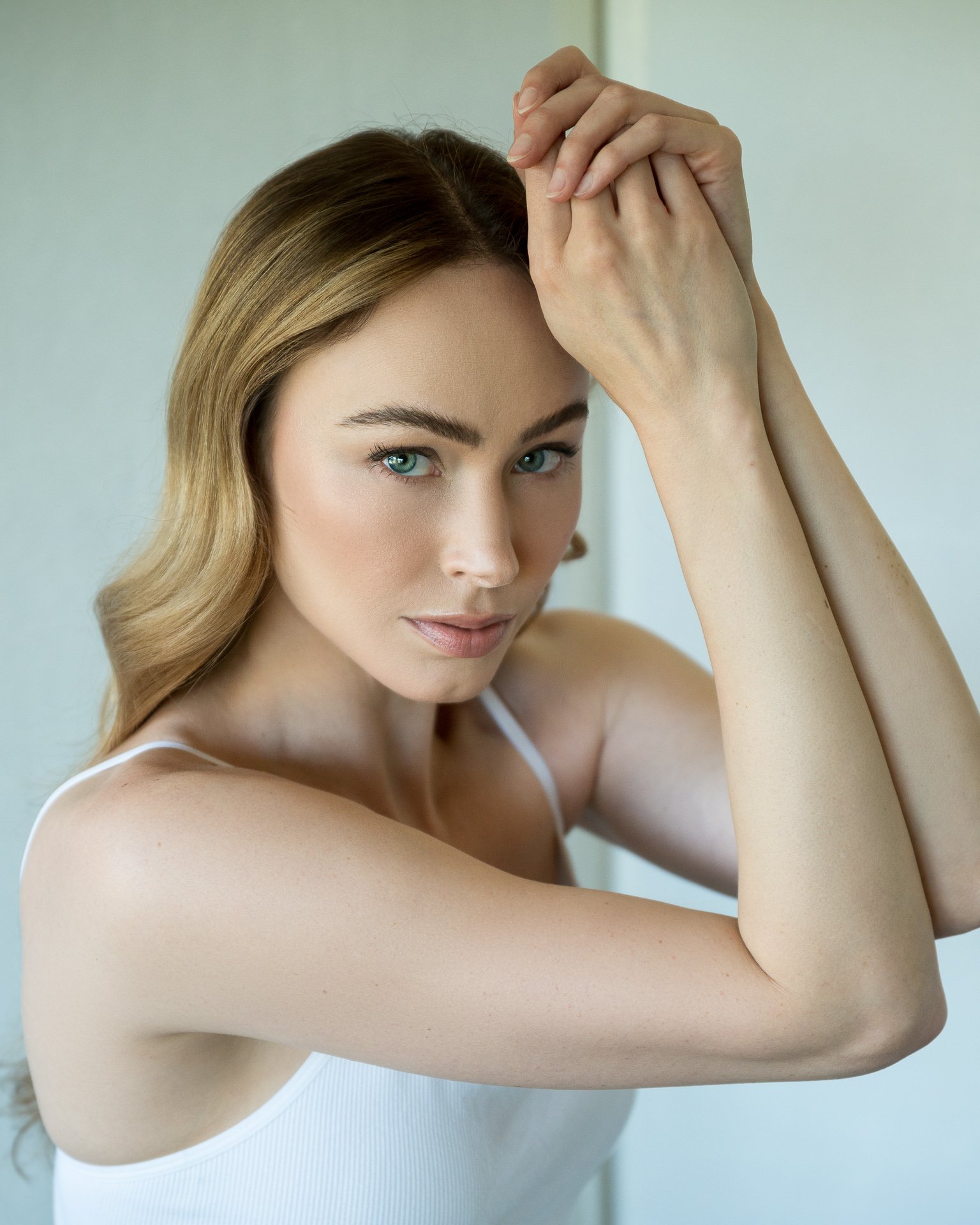A woman with light brown hair, blue eyes, and fair skin, wearing a white tank top, resting her head on her clasped hands, looking directly at the camera.