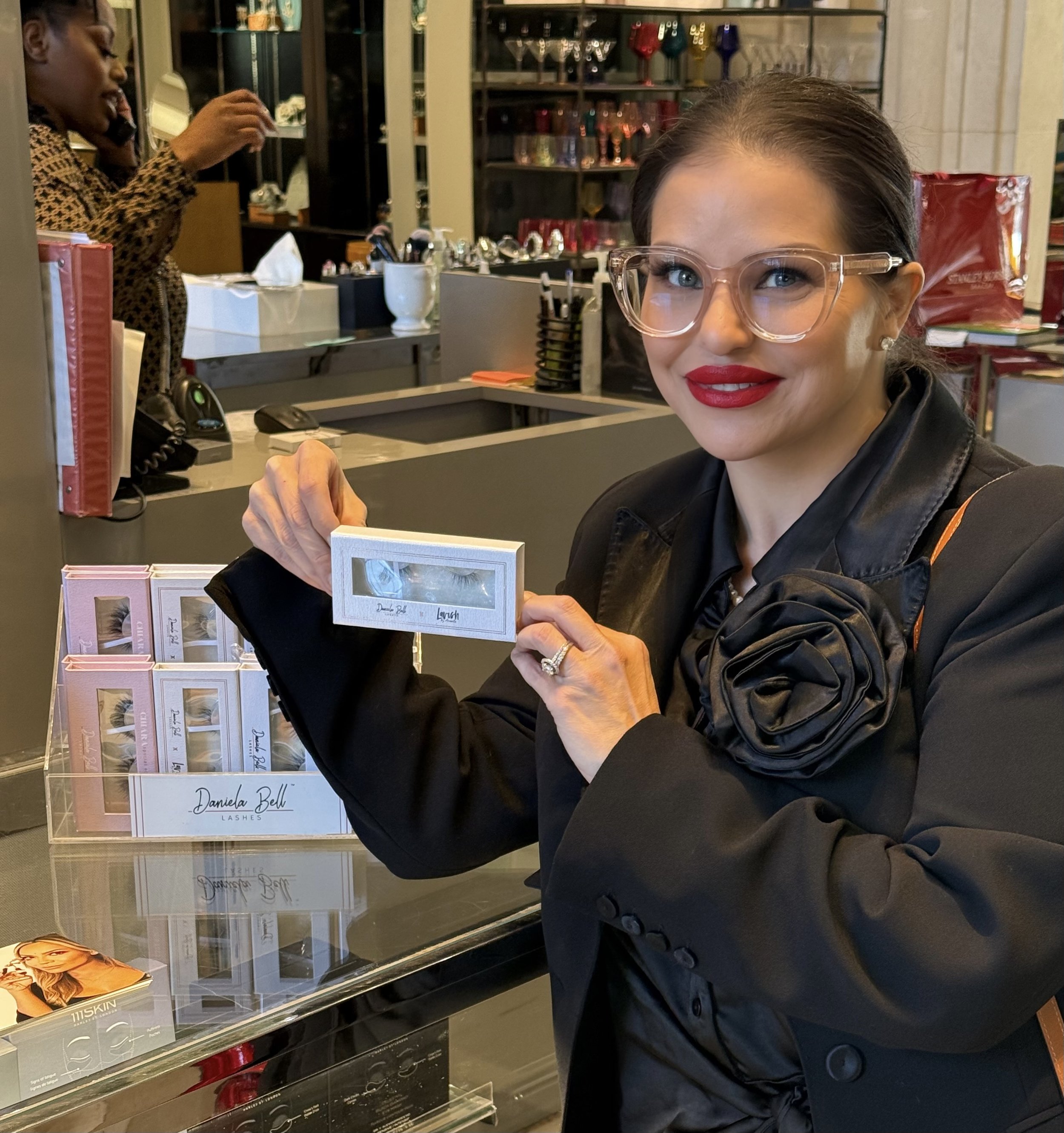 Woman with glasses and red lipstick holding a box of false eyelashes inside a beauty supply store.