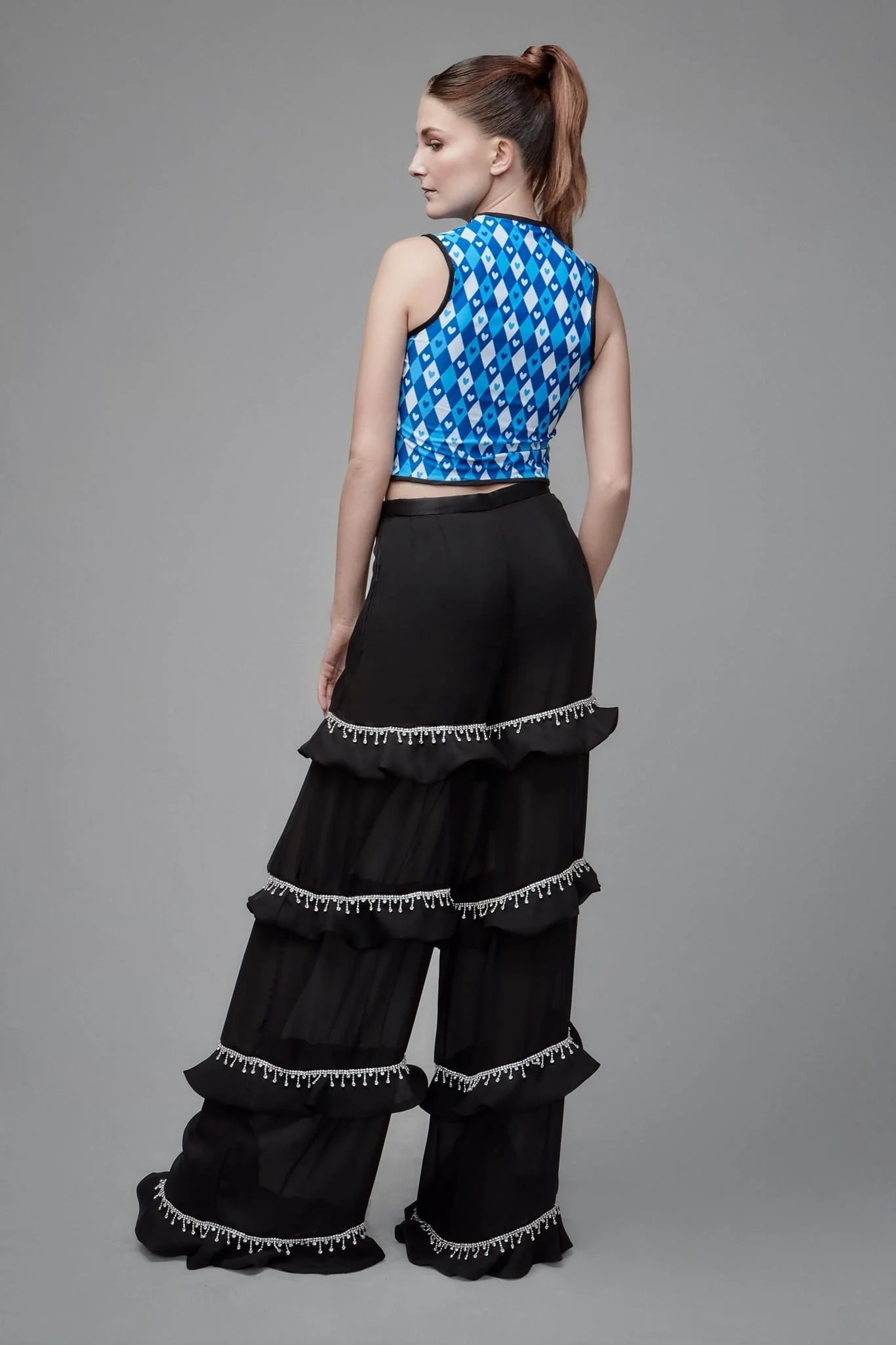 A young woman with long brown hair tied in a ponytail standing sideways against a plain gray background, wearing a sleeveless blue patterned crop top and black wide-leg pants with layered ruffled panels decorated with white fringe details.
