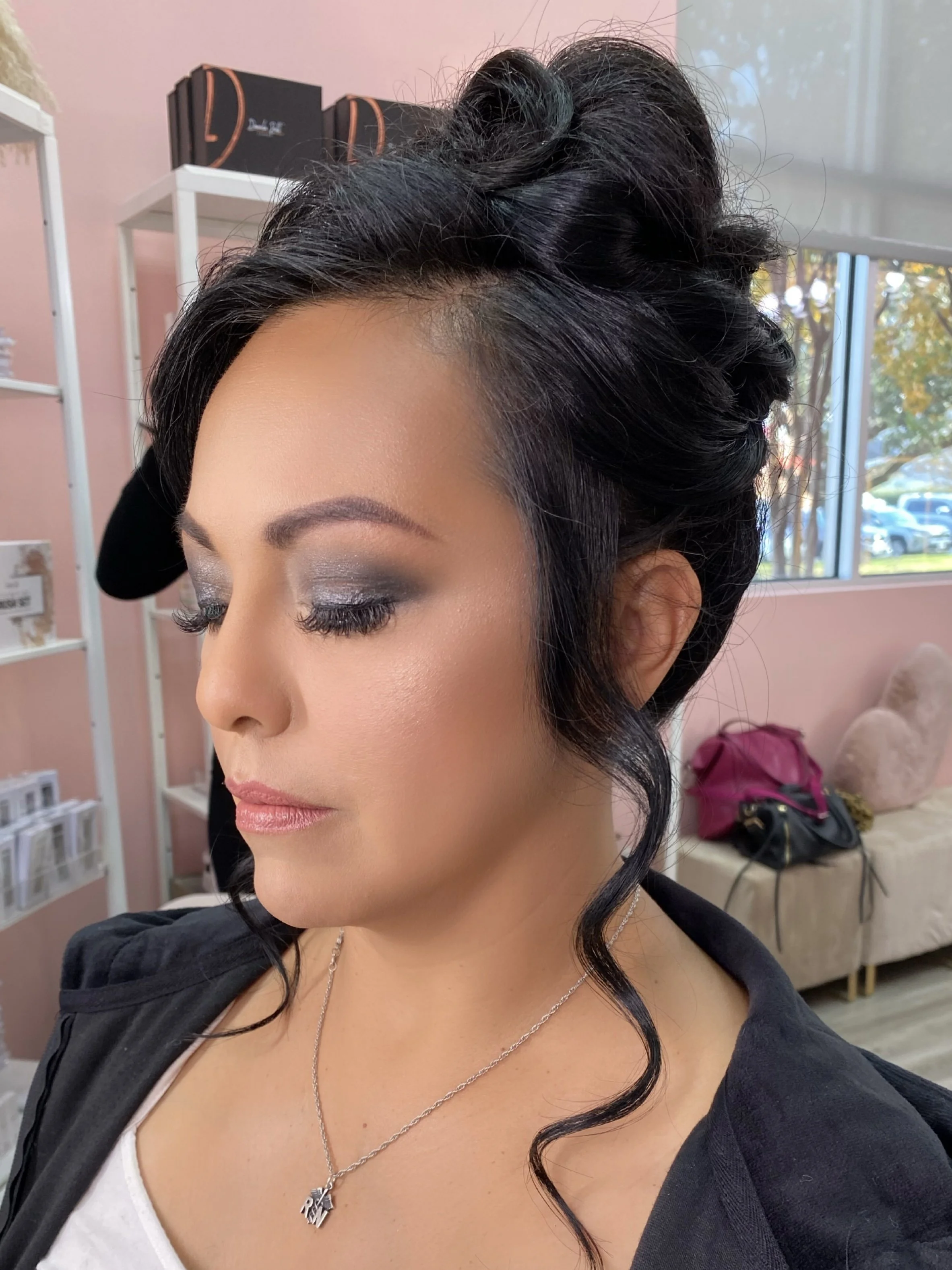 Close-up of a woman with styled black hair, makeup, and a necklace, indoors with pink walls and shelves in the background.