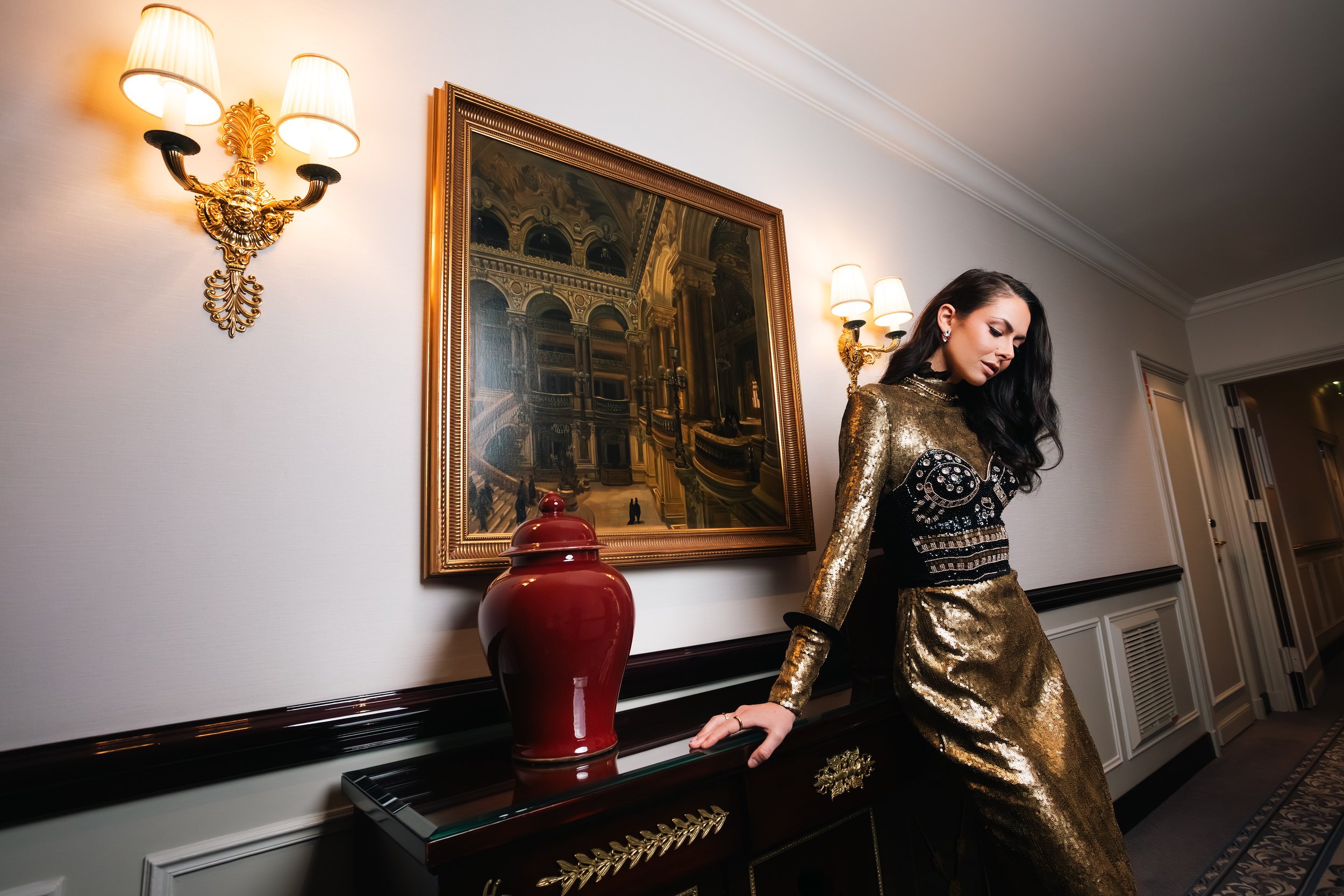 A woman in a gold sequined dress leaning on a black and gold decorated console table in an elegant hallway with a large framed mirror, red vase, and wall sconces.