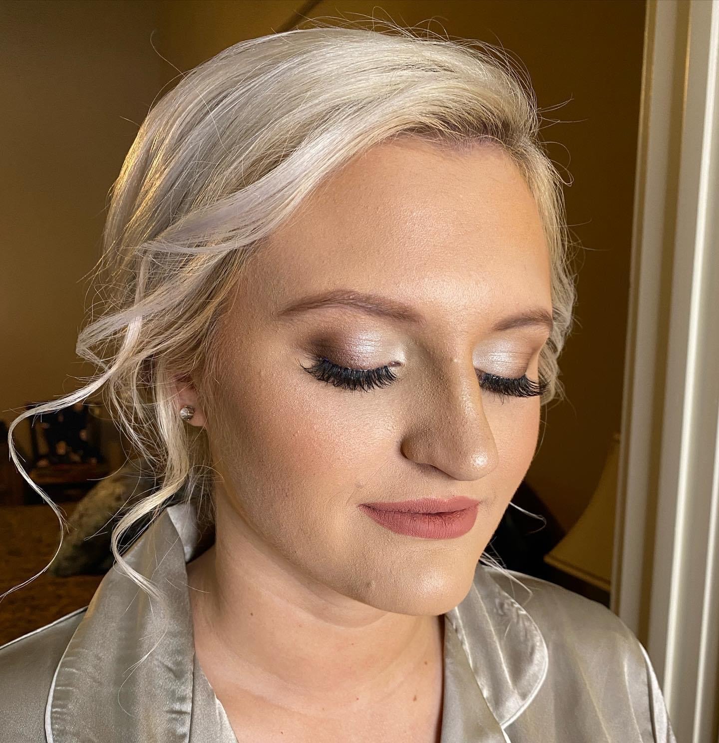 Close-up of a woman with platinum blonde hair styled in loose curls, wearing makeup with silver eyeshadow, long dark eyelashes, and matte lipstick. She is dressed in a silk robe and is in a softly lit room with neutral-colored walls.