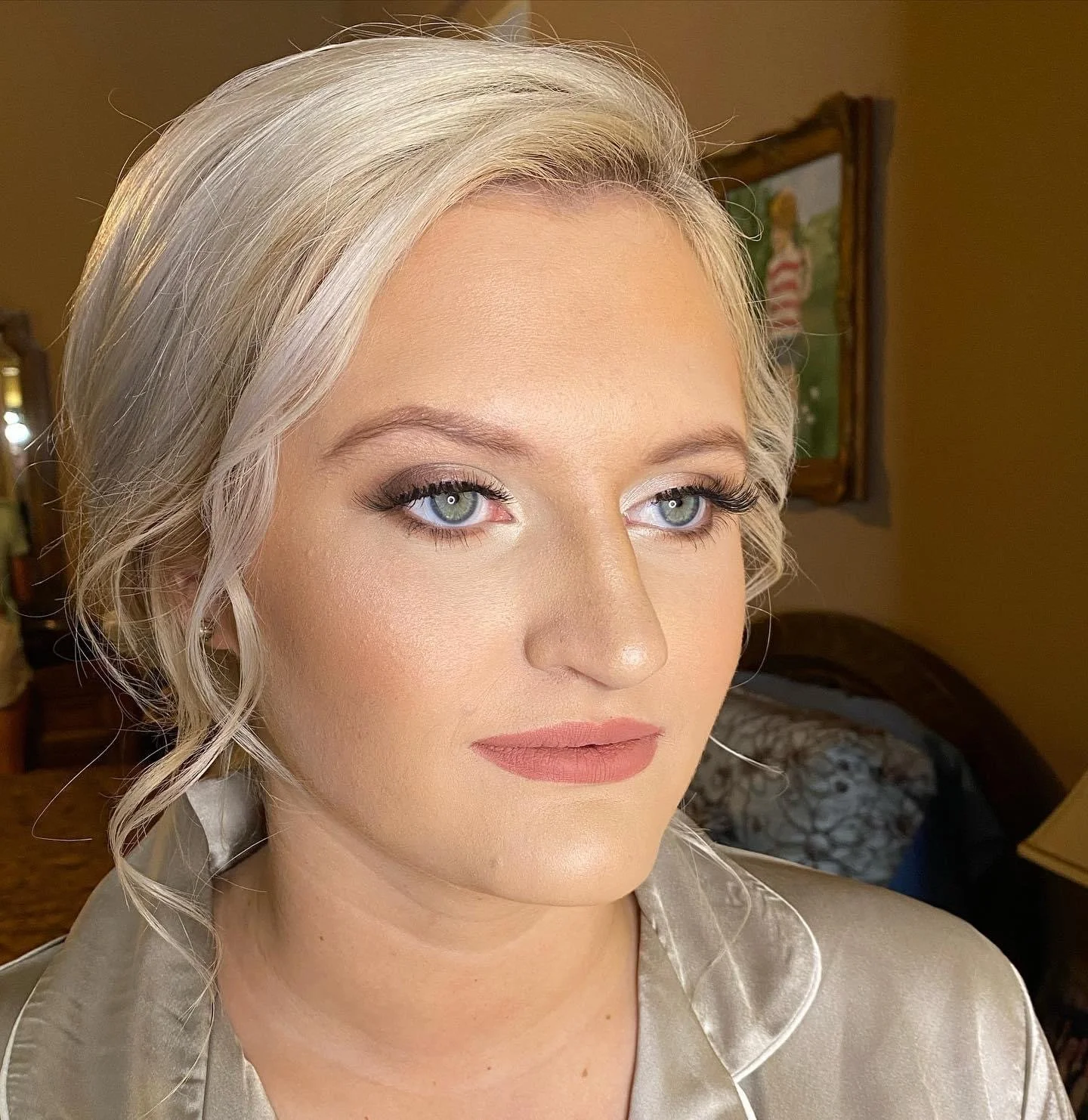 Close-up of a woman with short, wavy, blonde hair, blue eyes, wearing makeup, and a shiny beige silk robe, indoors with a painting on the wall behind her.