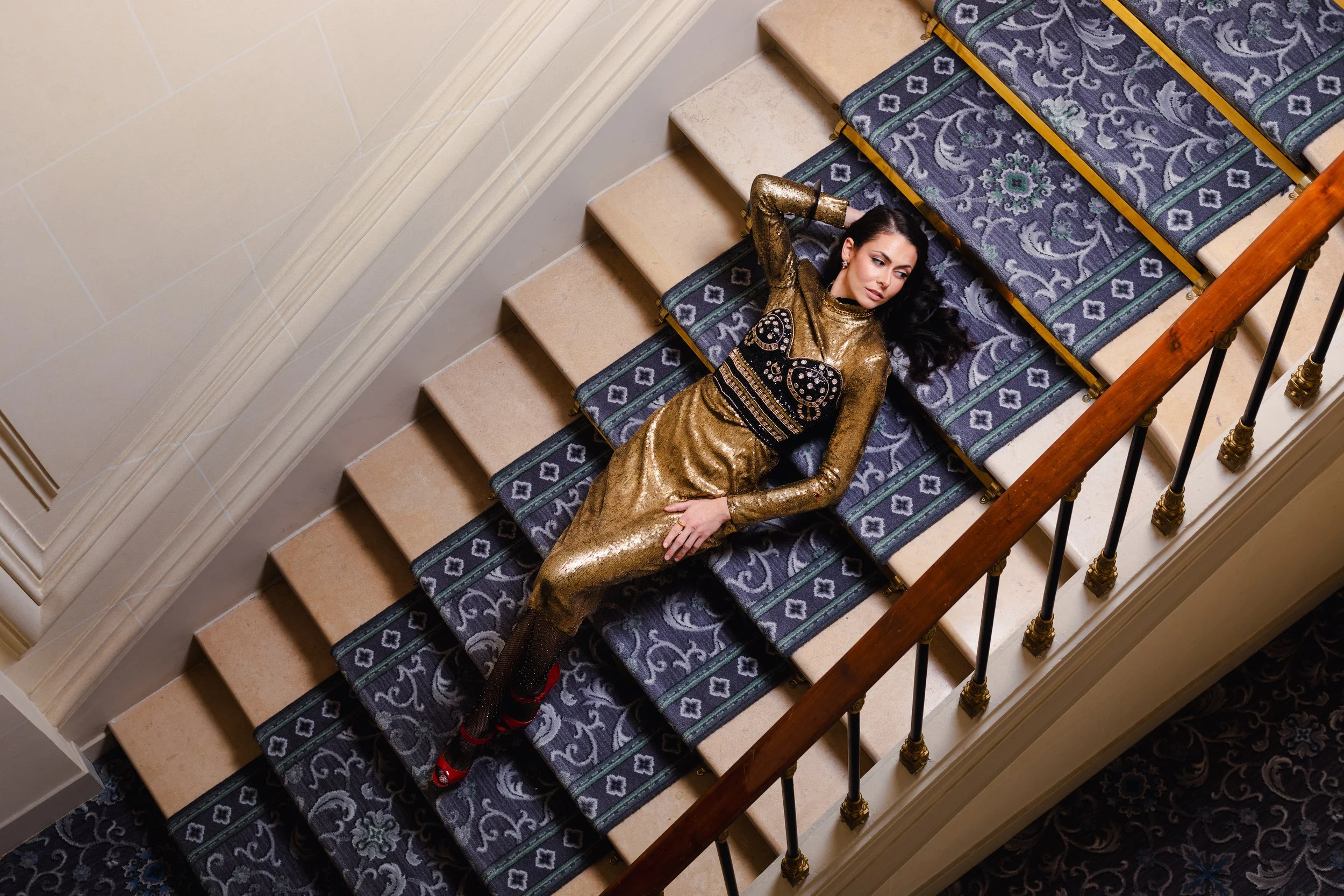 A woman lying on a staircase, wearing a shiny gold dress and black fishnet stockings, with red high heels, looking up with her hand on her head.