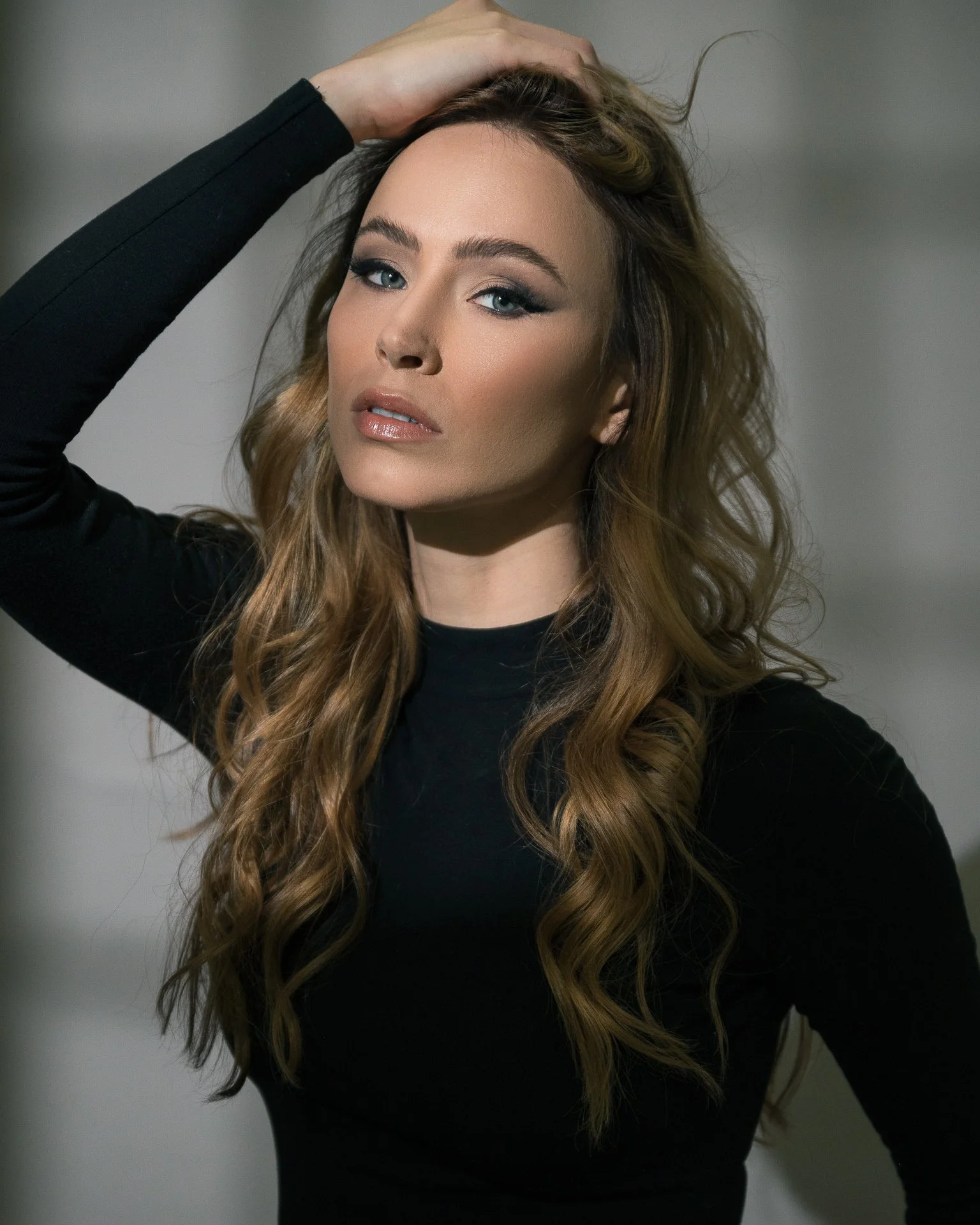 A woman with long wavy brown hair wearing a black top, posing with her hand on her head, looking directly at the camera.