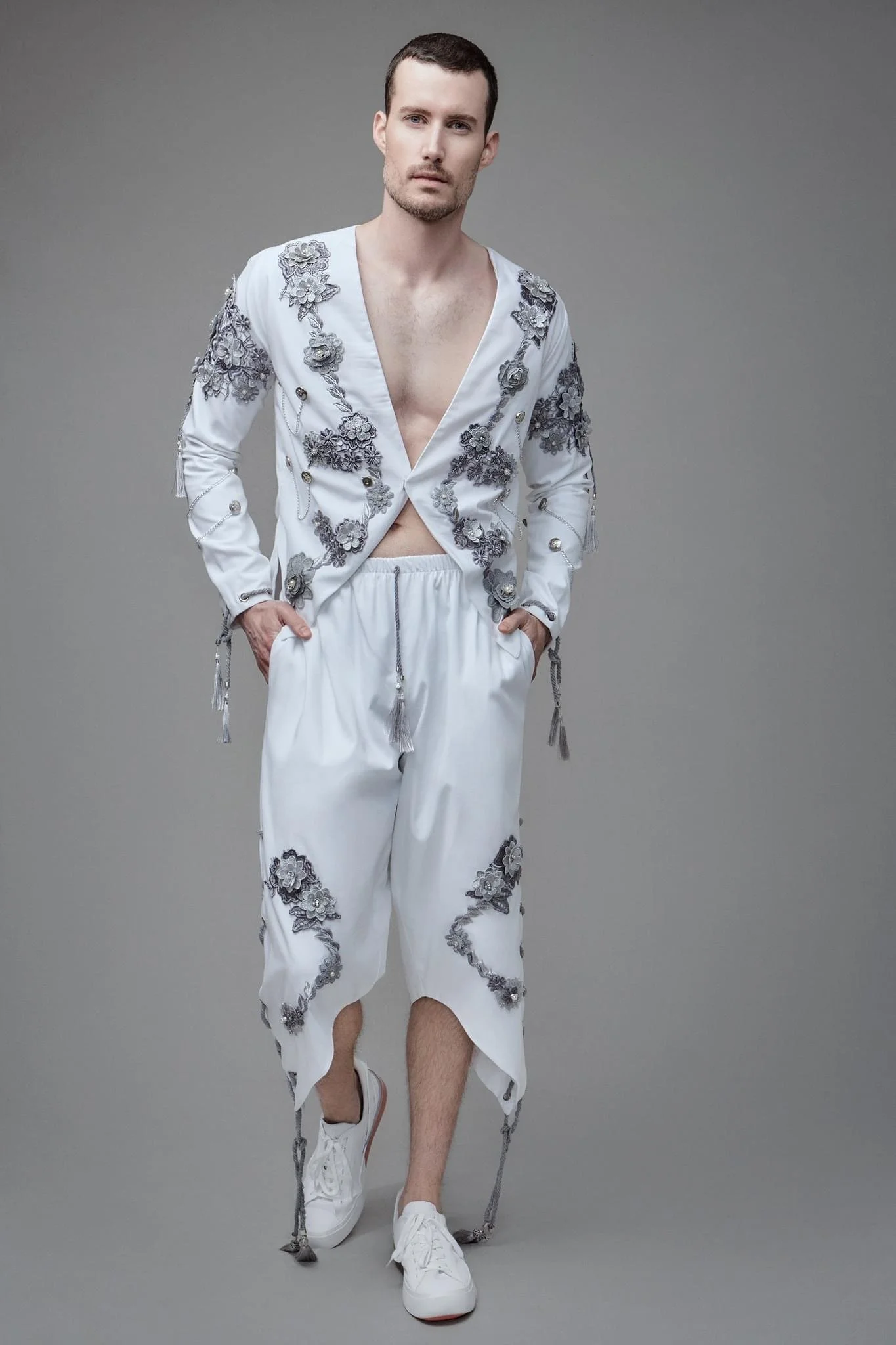 Man wearing a white embroidered jacket and matching pants with gray floral details, paired with white sneakers, standing against a gray background.