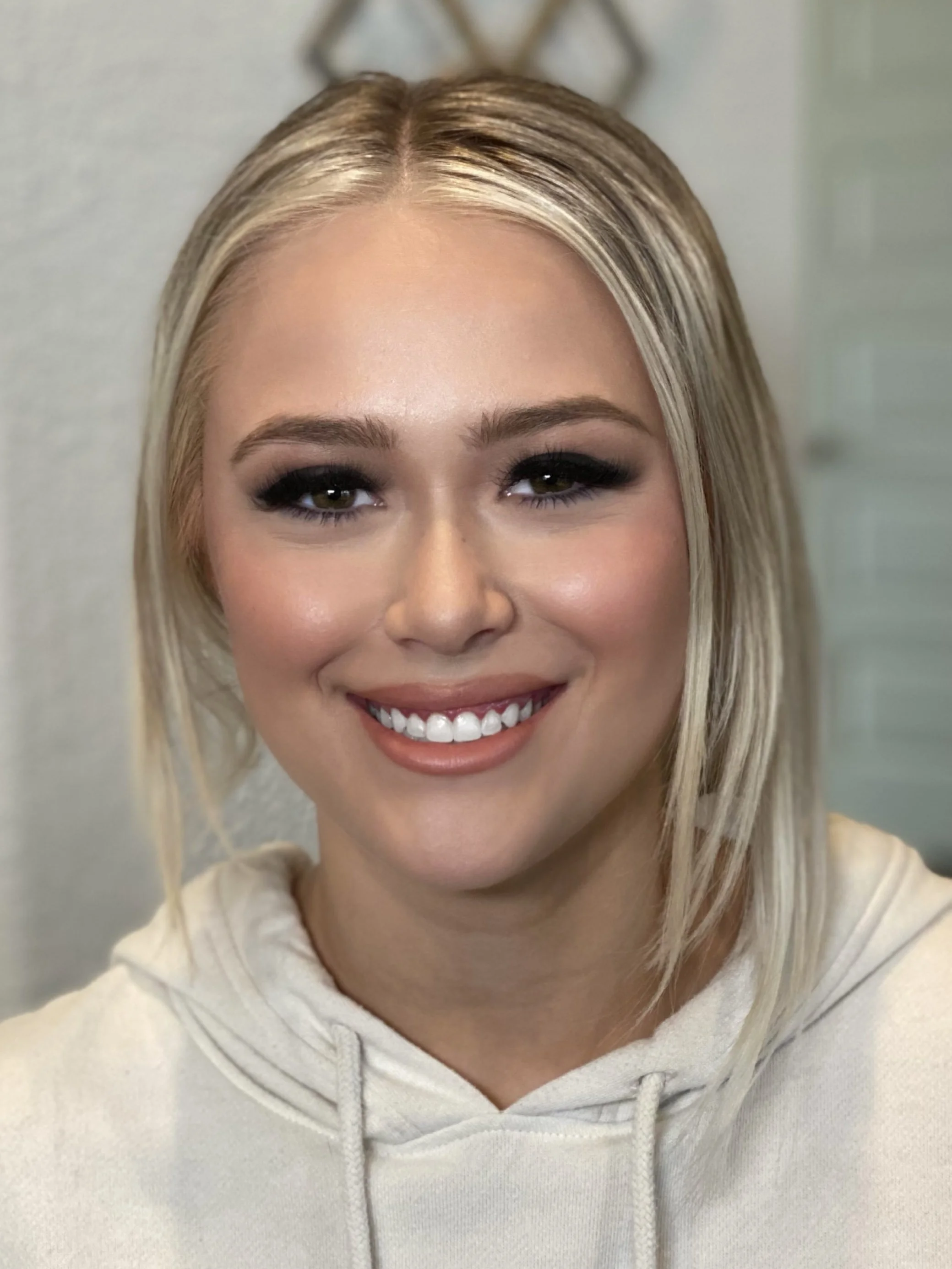 Close-up of a young woman with blonde hair, makeup, and a beige hoodie, smiling at the camera.