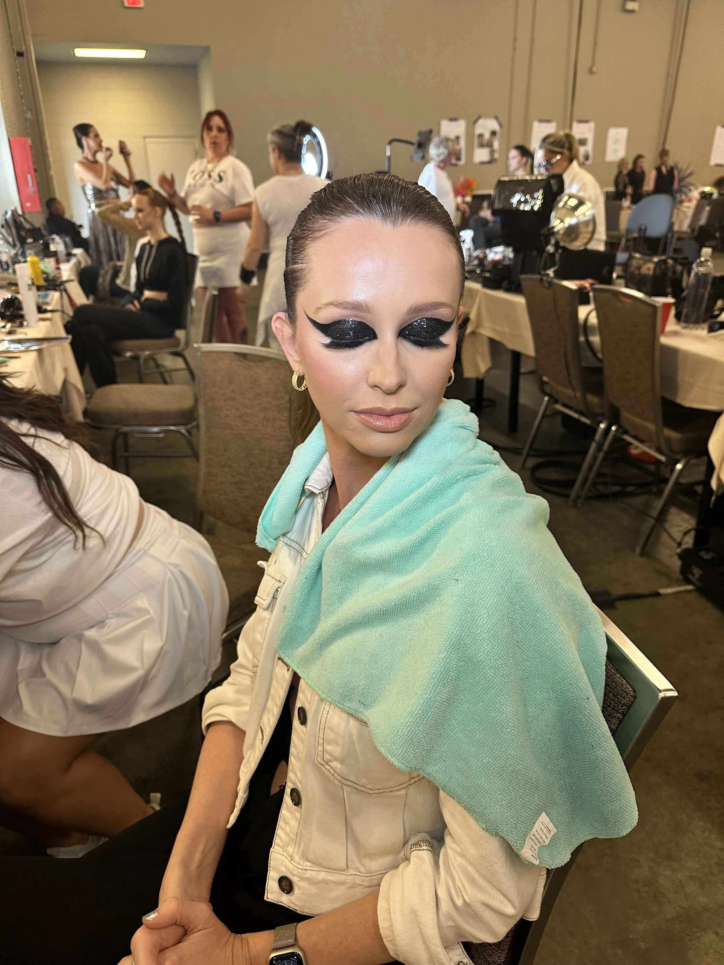 A woman with glamorous makeup and glittery black eyeshadow sitting with a light green towel draped over her shoulders in a busy behind-the-scenes makeup area.