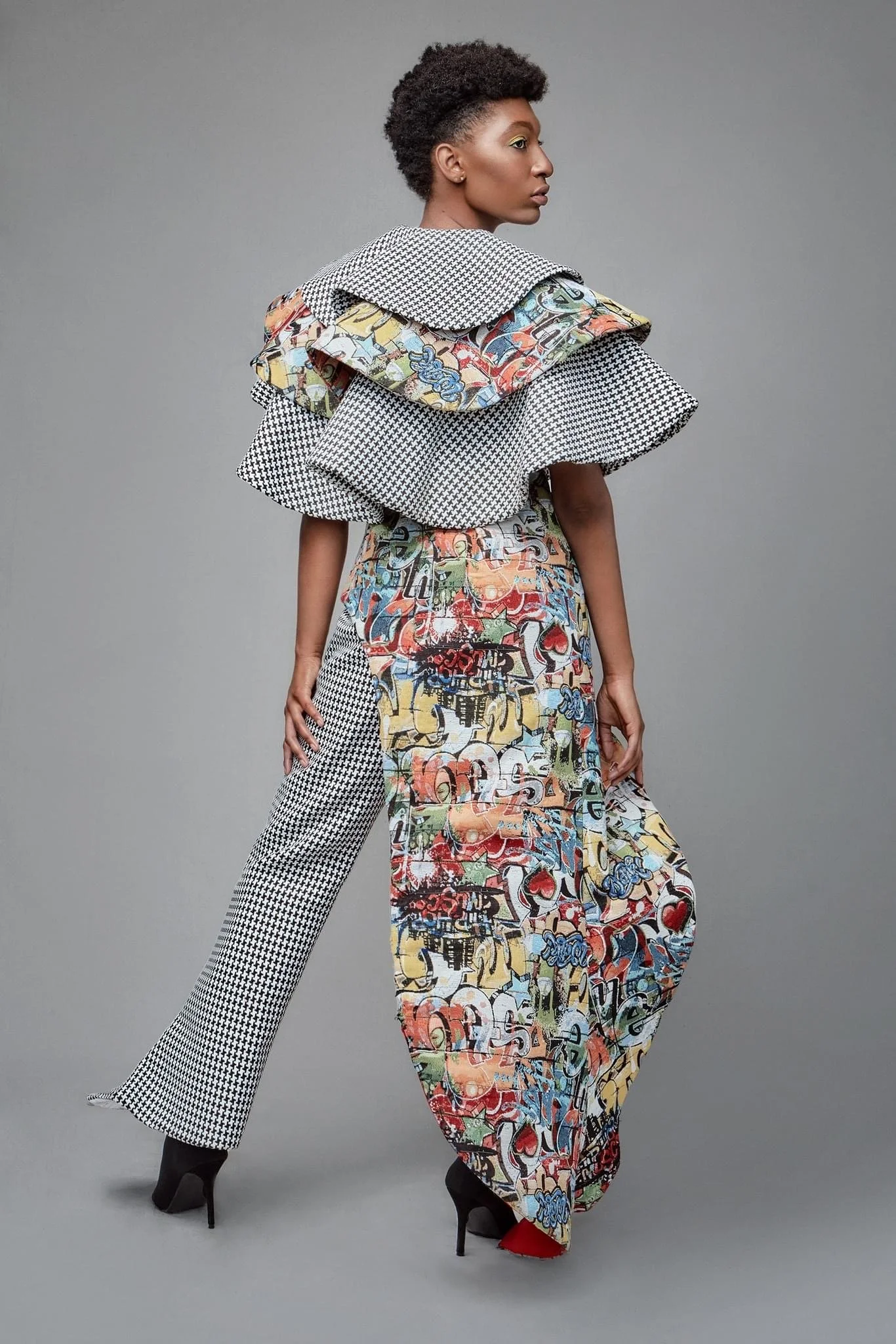 Woman wearing a colorful graffiti-patterned jumpsuit and layered black and white houndstooth fabric with black high heels against a gray background.