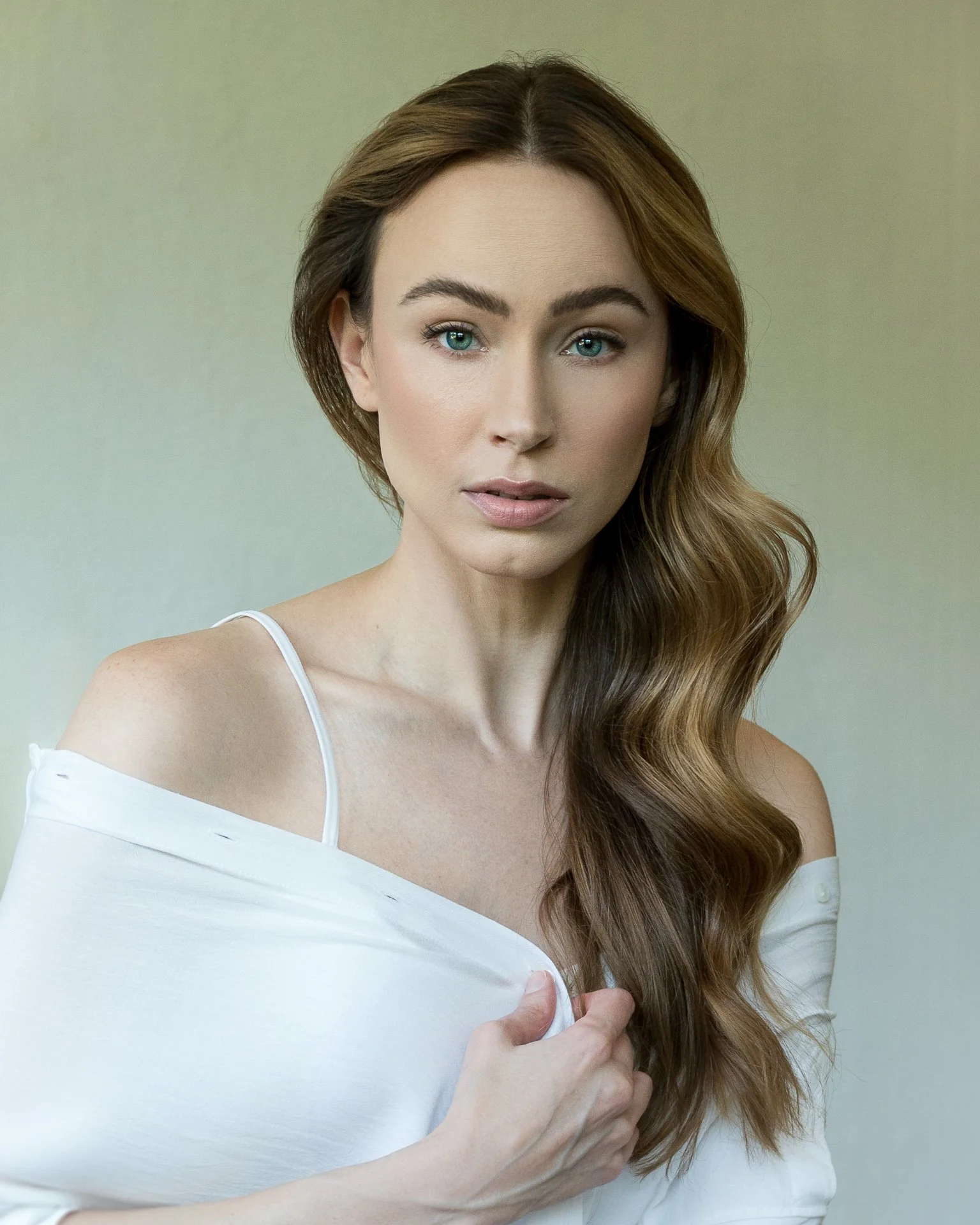 A woman with long wavy brown hair and blue eyes wearing a white off-shoulder top.