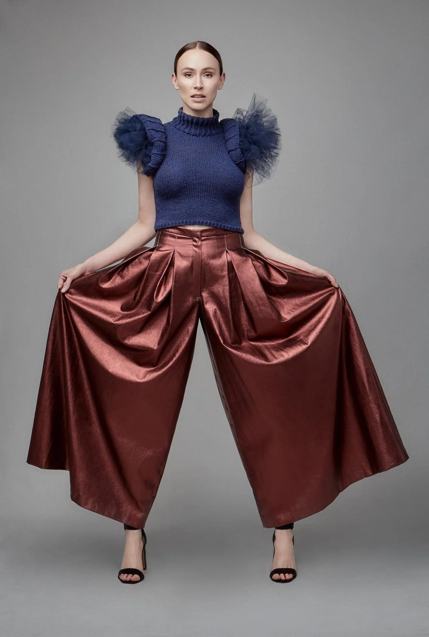 A woman wearing a blue knit top with puffed tulle sleeves, posing with her legs apart and holding the sides of her shiny, wide-legged red pants against a gray background.